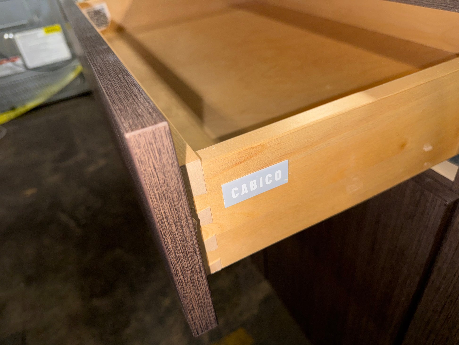 A close-up of a partially pulled-out wooden drawer with a dark brown wood-grain front and a light natural wood interior. A small white label on the drawer front reads CABICO.