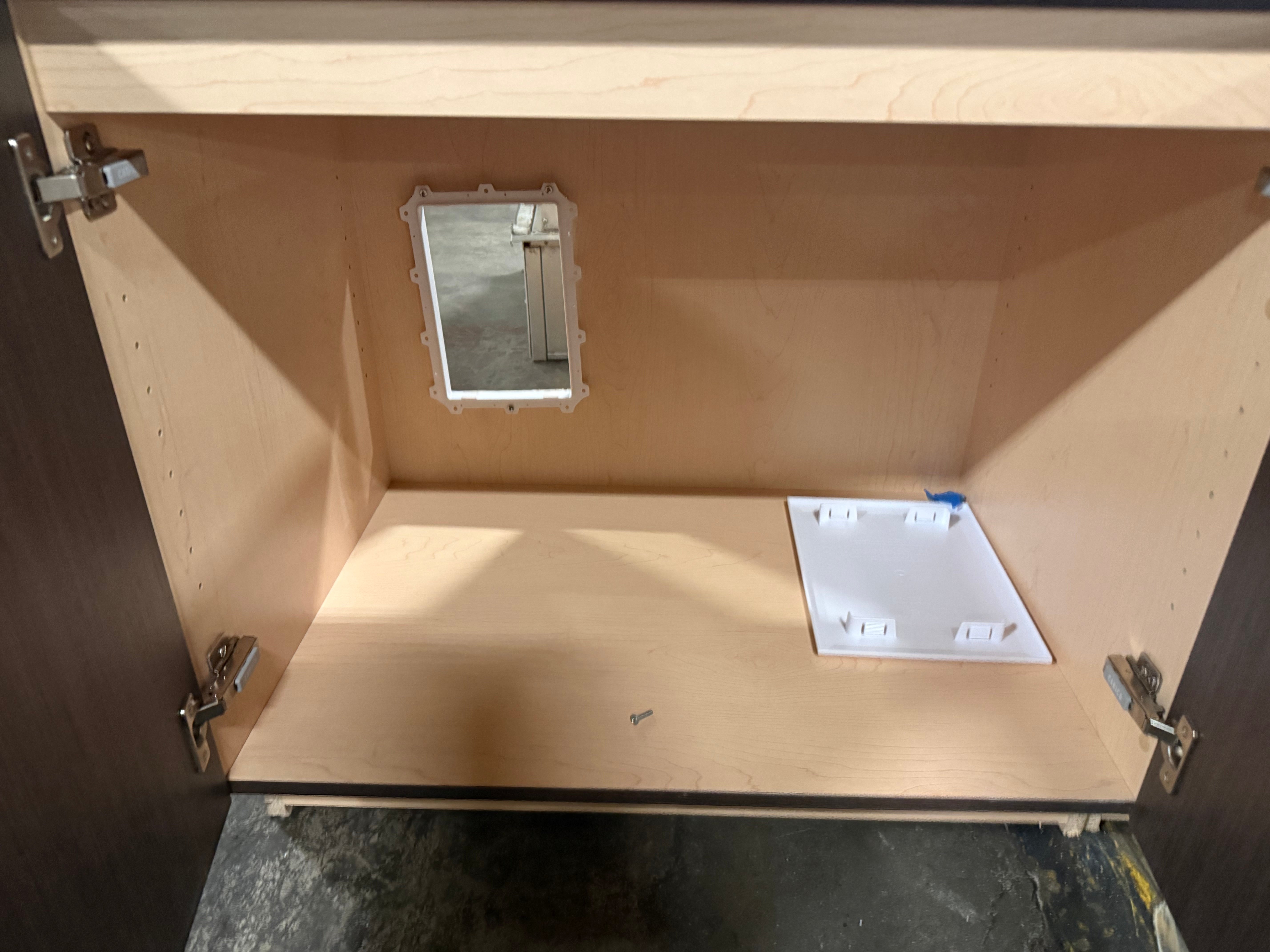An open wooden cabinet interior with light natural wood panels, a small square mirror mounted on the back wall, and a white rectangular accessory or tray on the bottom shelf. The cabinet is photographed in a workshop or storage setting with dark floor visible.