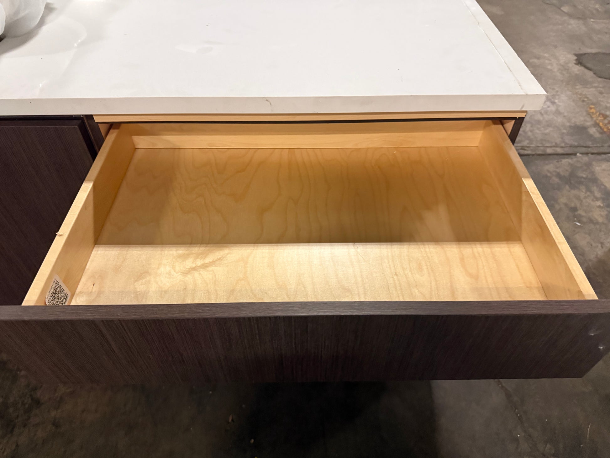 A close-up of a dark wood drawer with its front panel pulled open, showing a light natural wood interior, set beneath a white countertop.