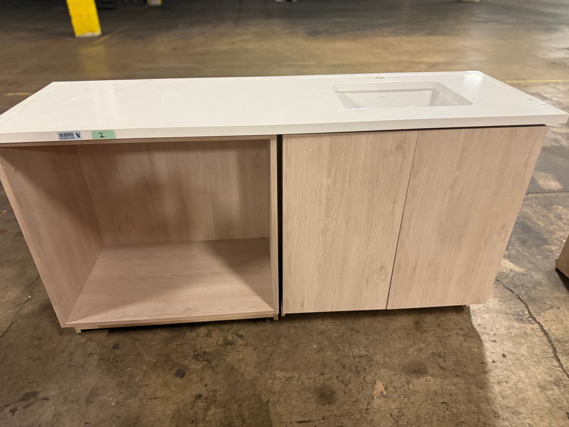 A light wood kitchen island cabinet with a white countertop and an integrated sink, photographed in a warehouse setting on a concrete floor.