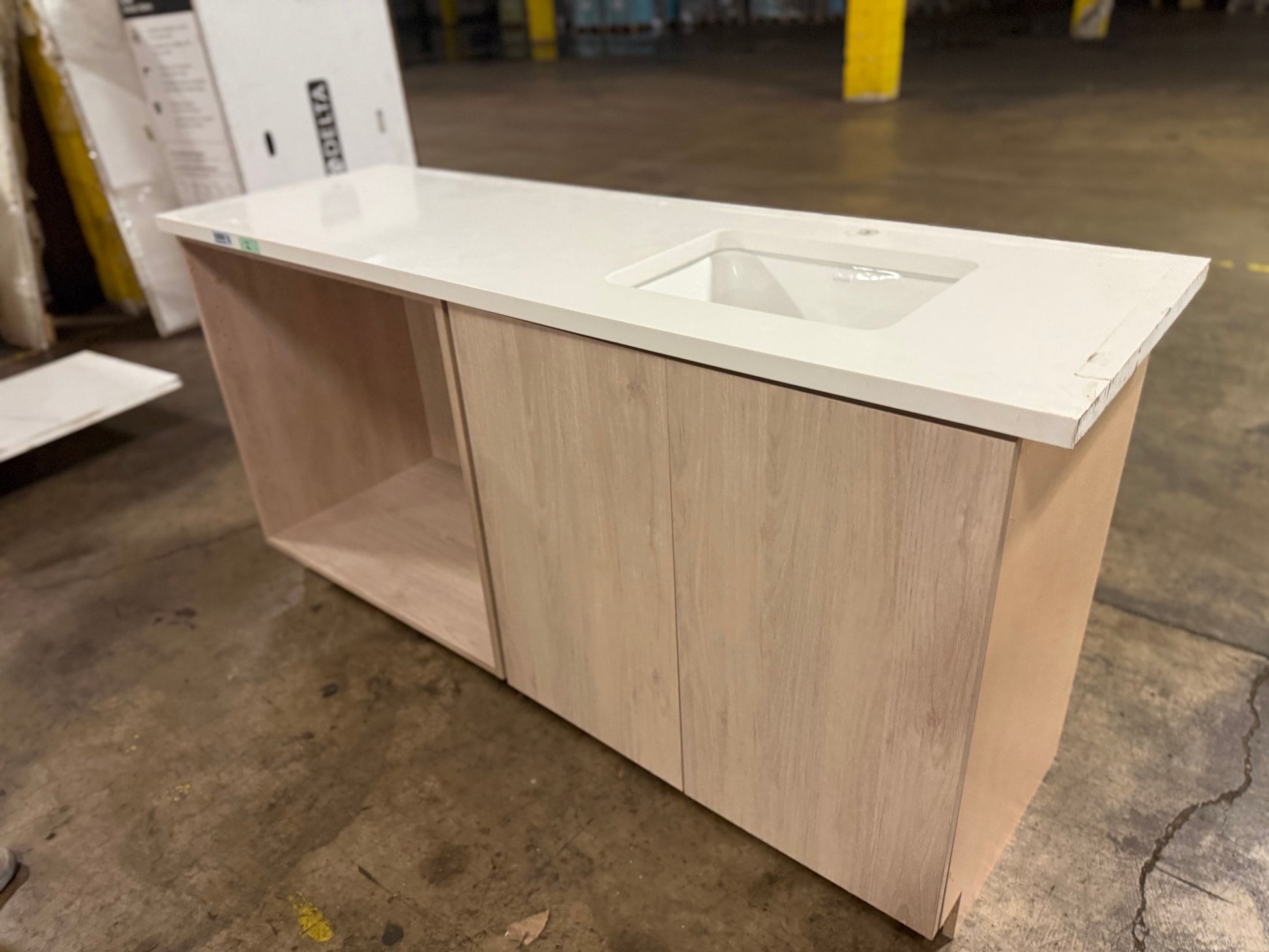 A light wood vanity cabinet with a white countertop and integrated sink, photographed in a warehouse setting on a concrete floor.