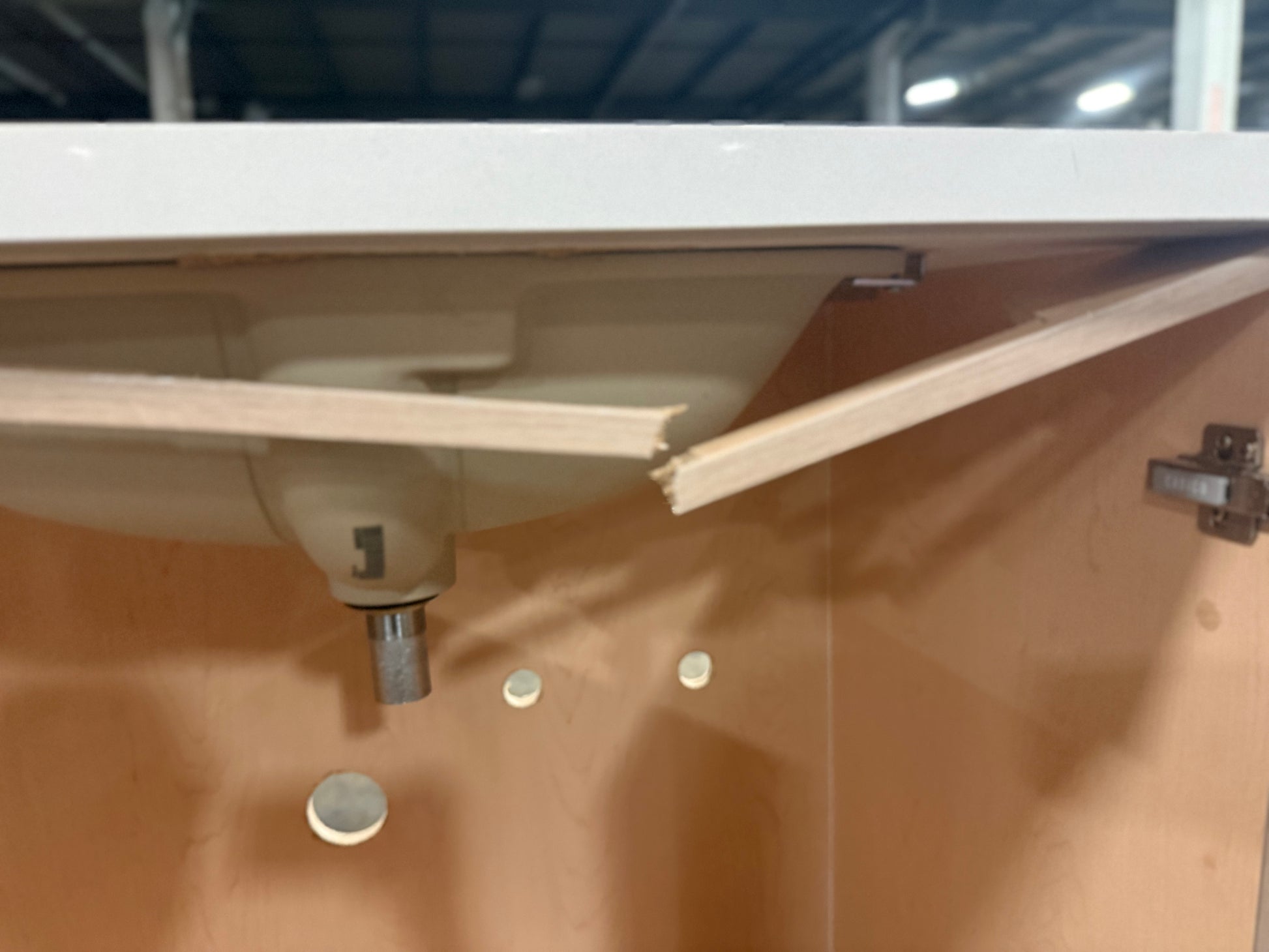Close-up of a kitchen cabinet interior with a sink mounted underneath a white countertop. The cabinet door is open, showing a light wood interior, metal hinges, and a sink base with a visible drain pipe.