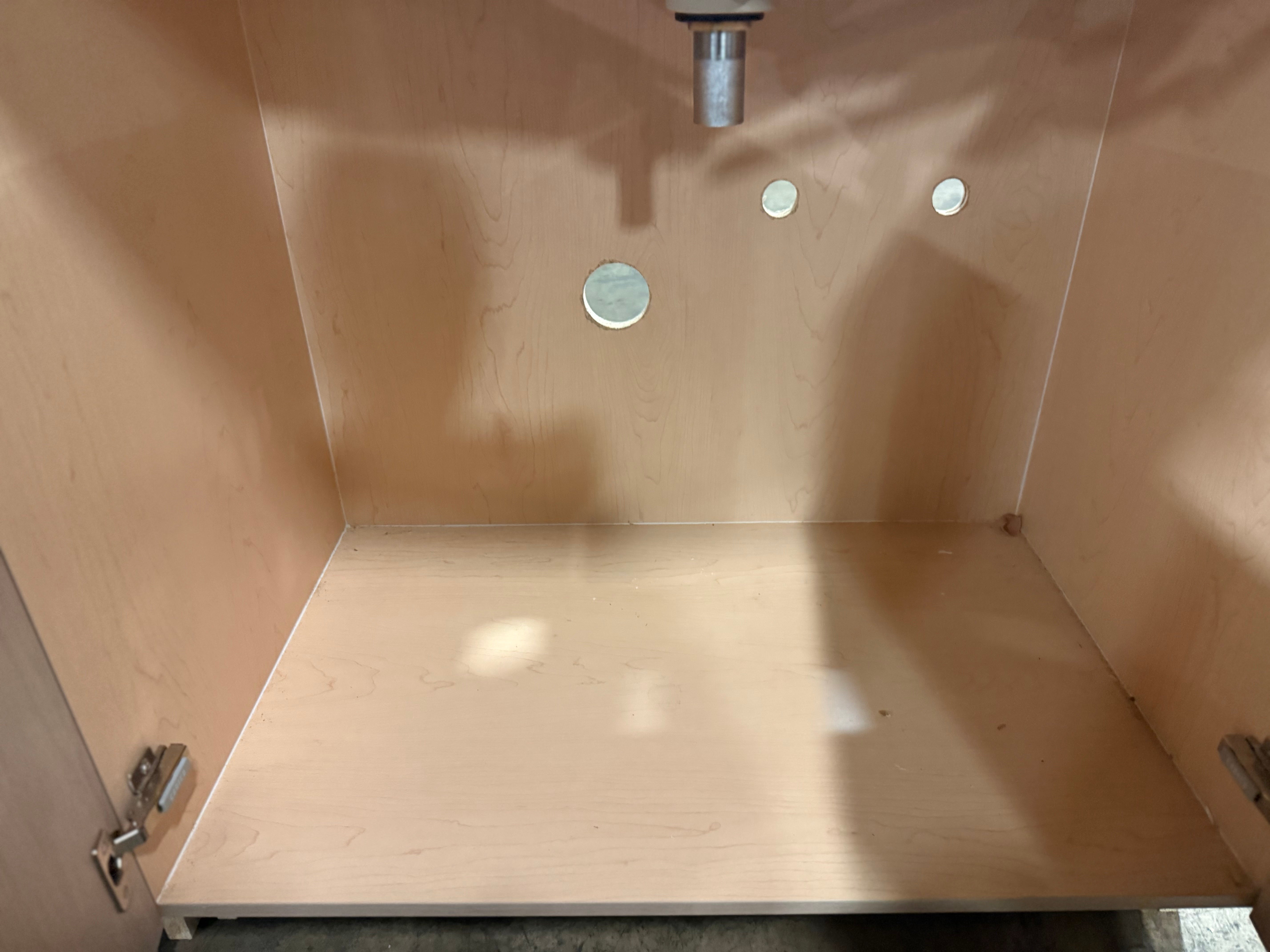 An empty light wood cabinet interior with a plywood base, back panel, and side panels, showing metal plumbing hardware and a drain pipe at the top.