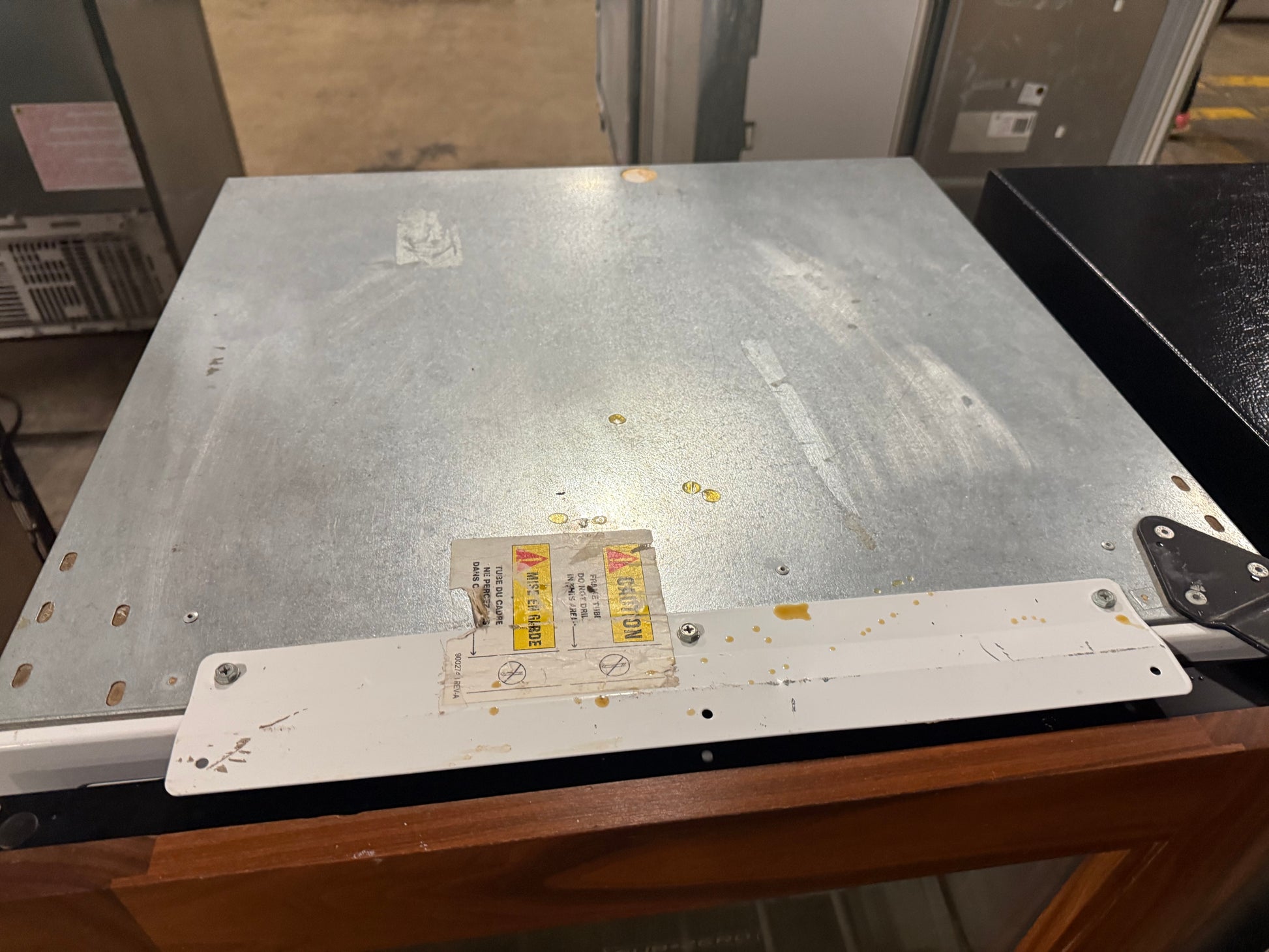 A large silver metal server rack module or industrial panel with a white mounting bracket and stickers, photographed on a wooden table in a warehouse or equipment room.