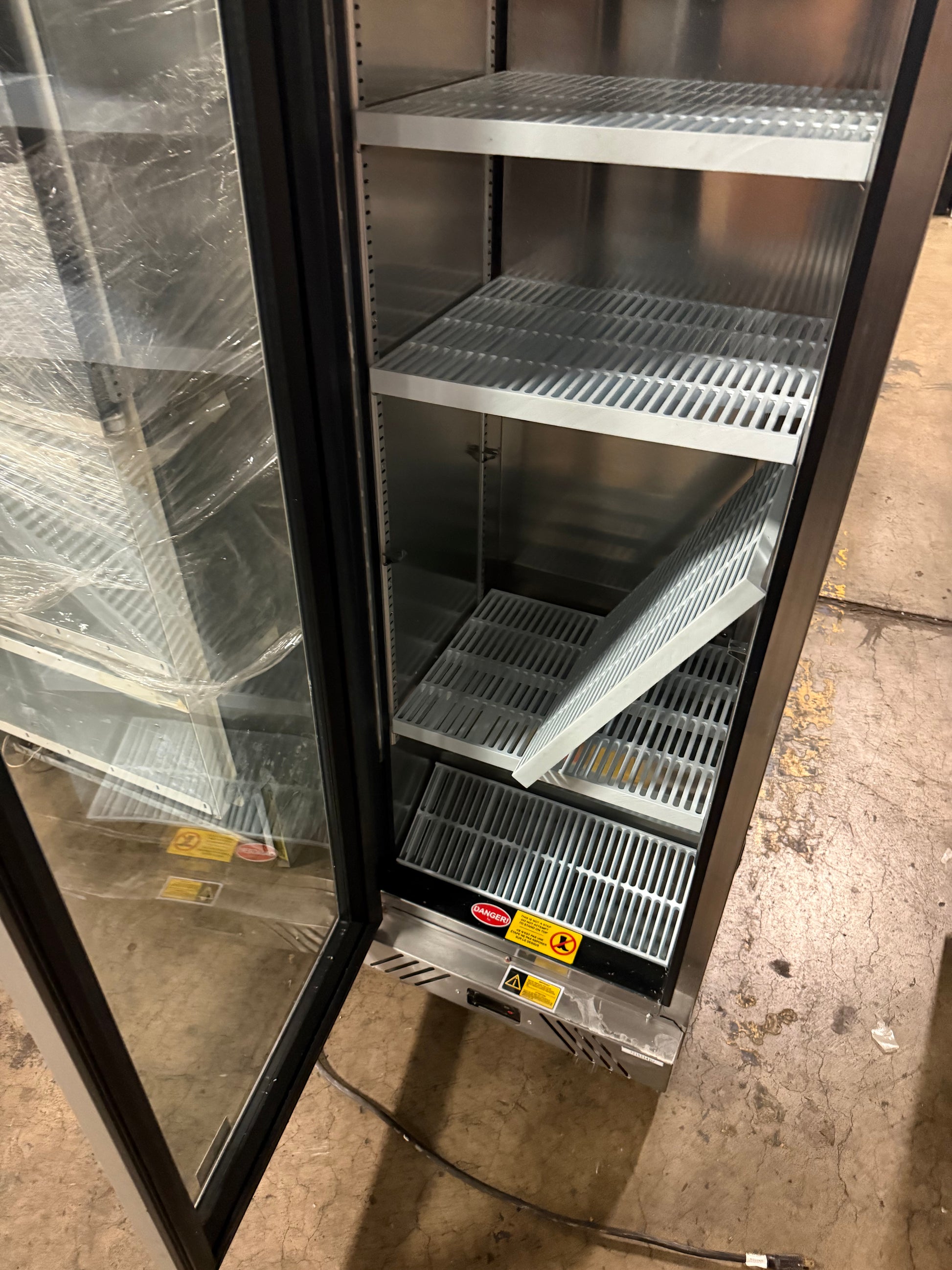 A stainless steel commercial glass-door refrigerator with the door open, showing three empty metal shelves inside. The unit is photographed on a concrete floor in a warehouse or retail setting.