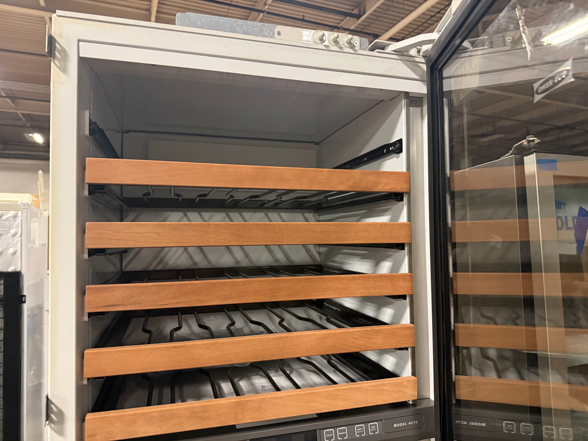 A wine cooler or refrigerator with its door open, showing multiple wooden shelves and a black wire rack inside. The appliance has a white exterior and a glass door, with another similar unit partially visible on the right.