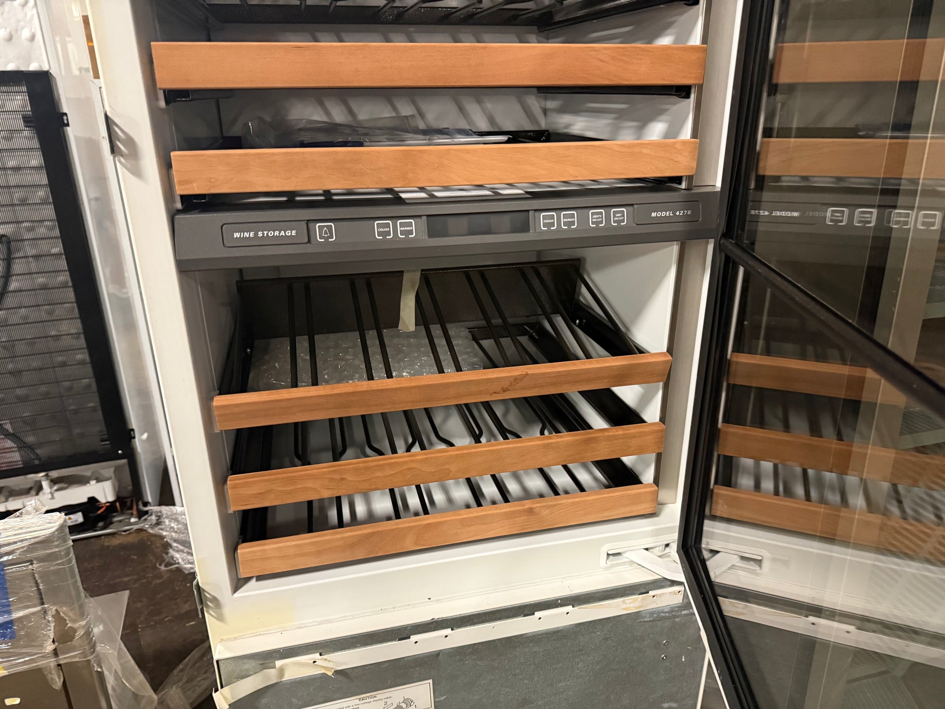 A wine refrigerator with multiple shelves, featuring light wood slat fronts, a dark control panel, and glass doors, shown in what appears to be a showroom or warehouse setting.