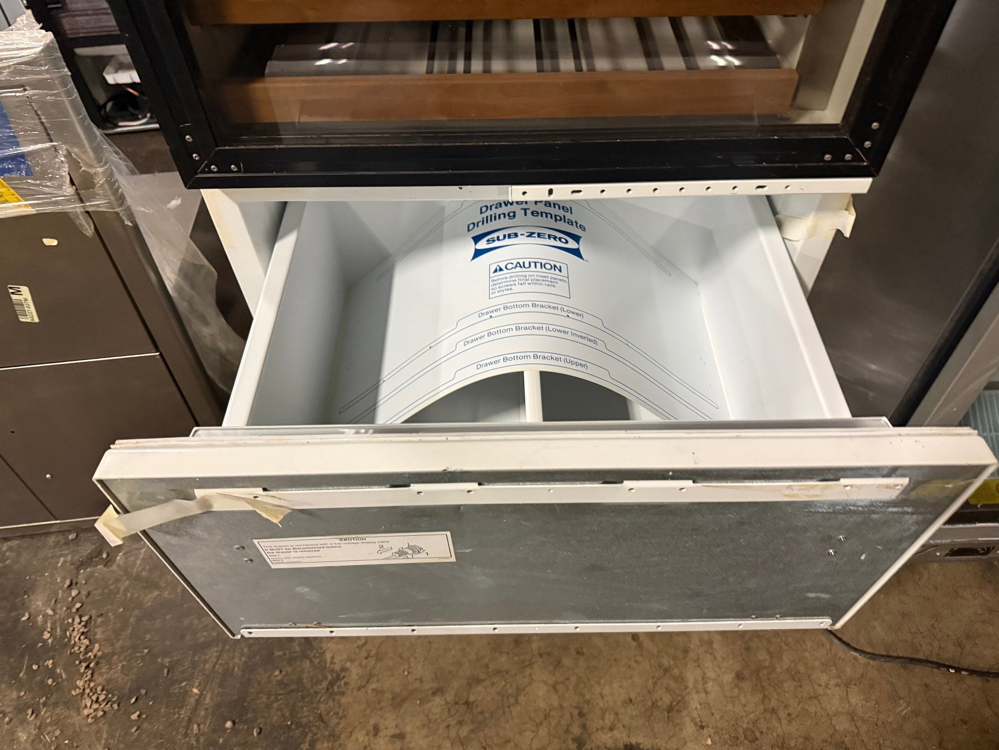 An open white and silver refrigerator door showing the interior shelves and a paper liner inside, photographed in a warehouse or storage area.