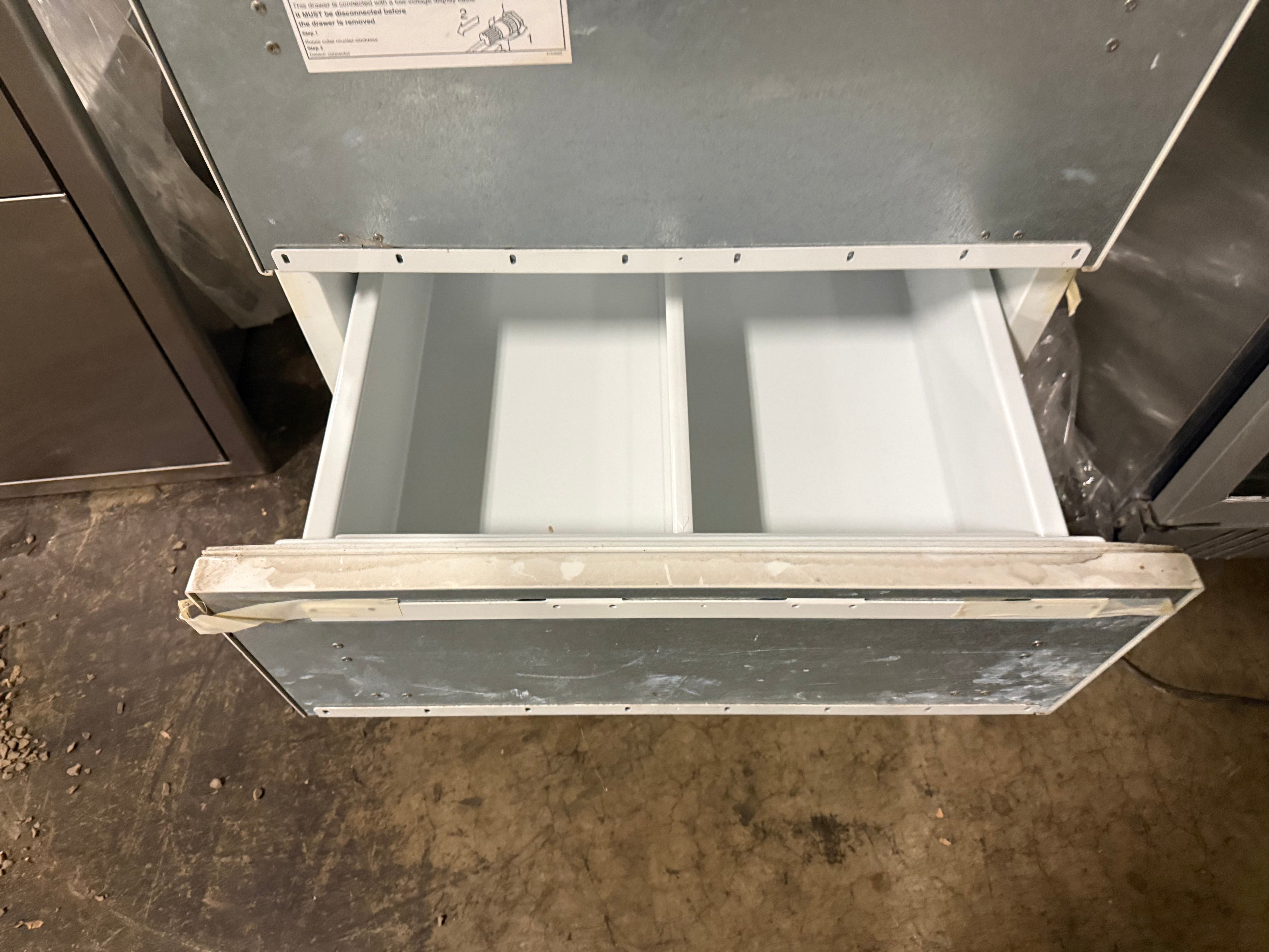 A stainless steel commercial refrigerator drawer pulled open, showing a two-compartment interior, set on a concrete floor.