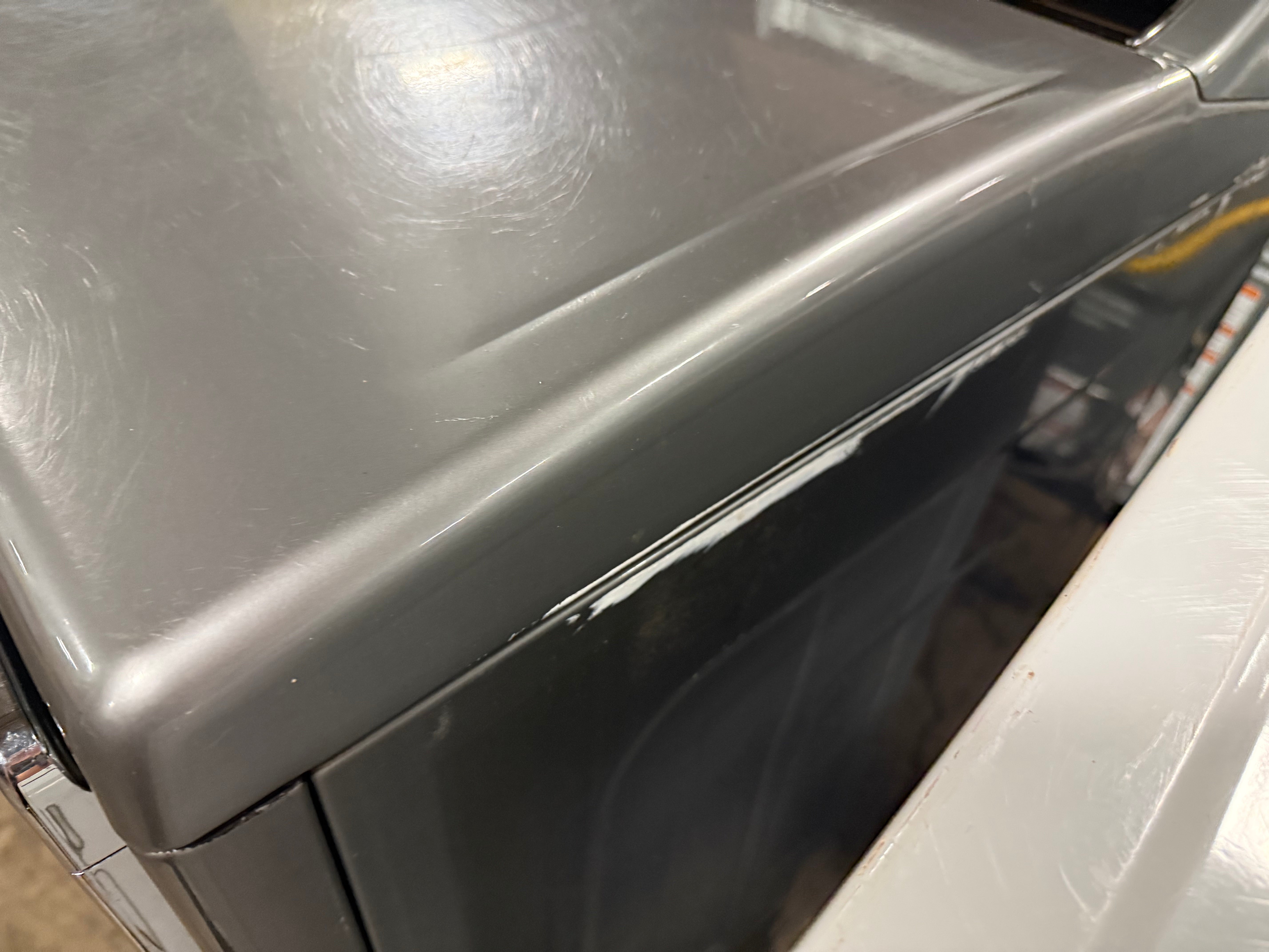 Close-up of a gray appliance, likely a washing machine or dryer, showing the top panel with visible scratches and scuffs, alongside a white unit next to it.