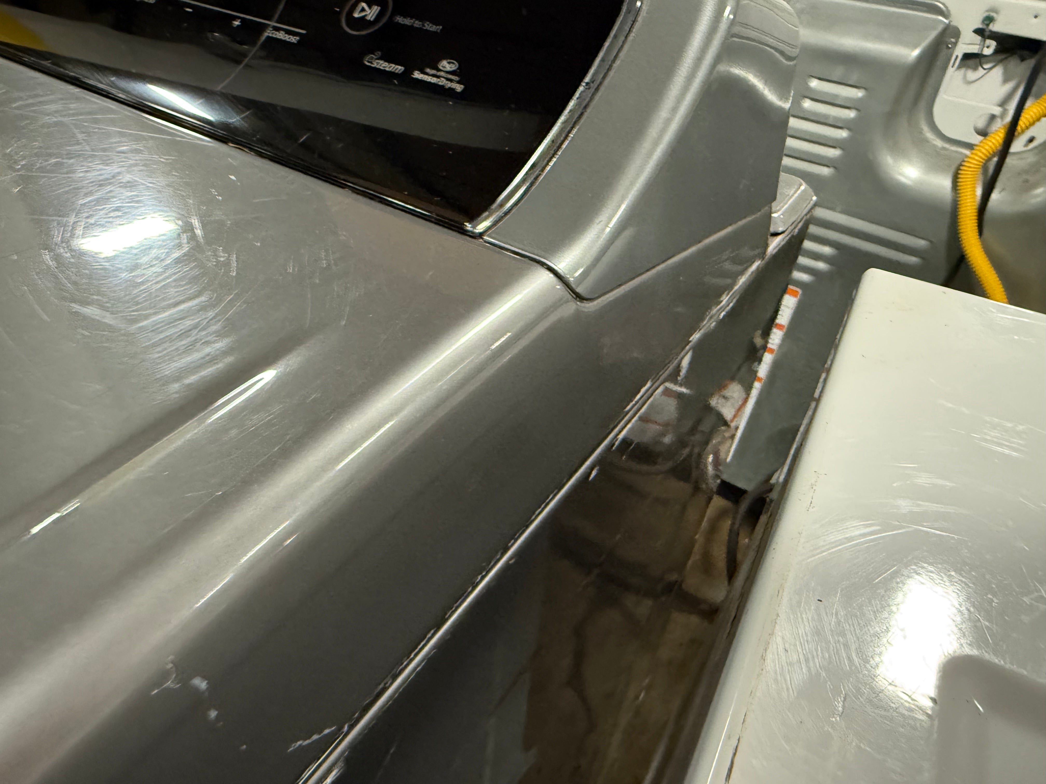 A close-up view of a gray appliance, likely a washing machine or dryer, showing the side panel, control console, and adjacent white unit in a utility room setting.