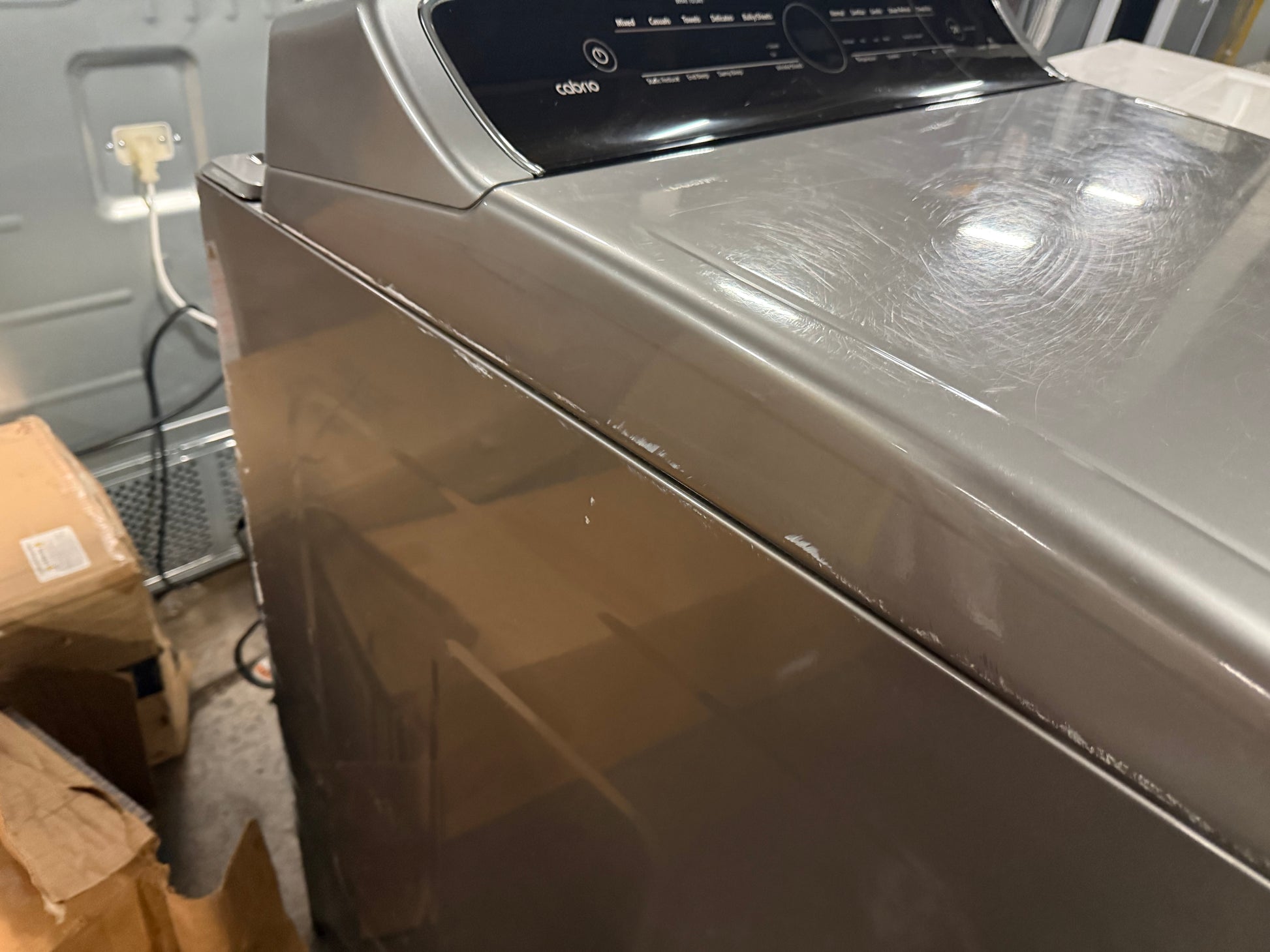 A top-loading clothes washer in a metallic taupe-gray finish, photographed in a warehouse or storage setting with cardboard boxes and an electrical outlet visible in the background.