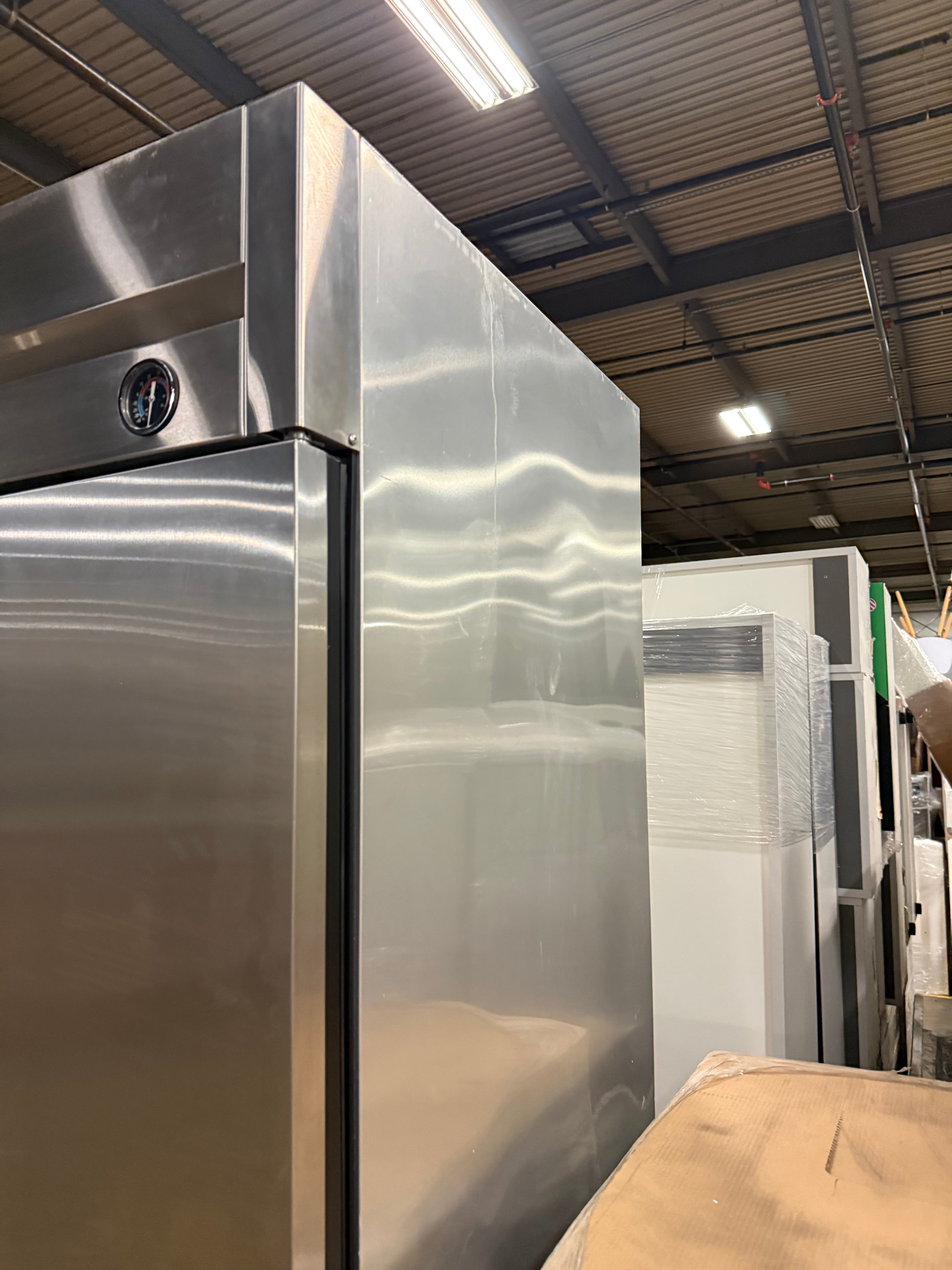 A large stainless steel commercial refrigerator standing in a warehouse or showroom, with other appliances and ceiling lights visible in the background.