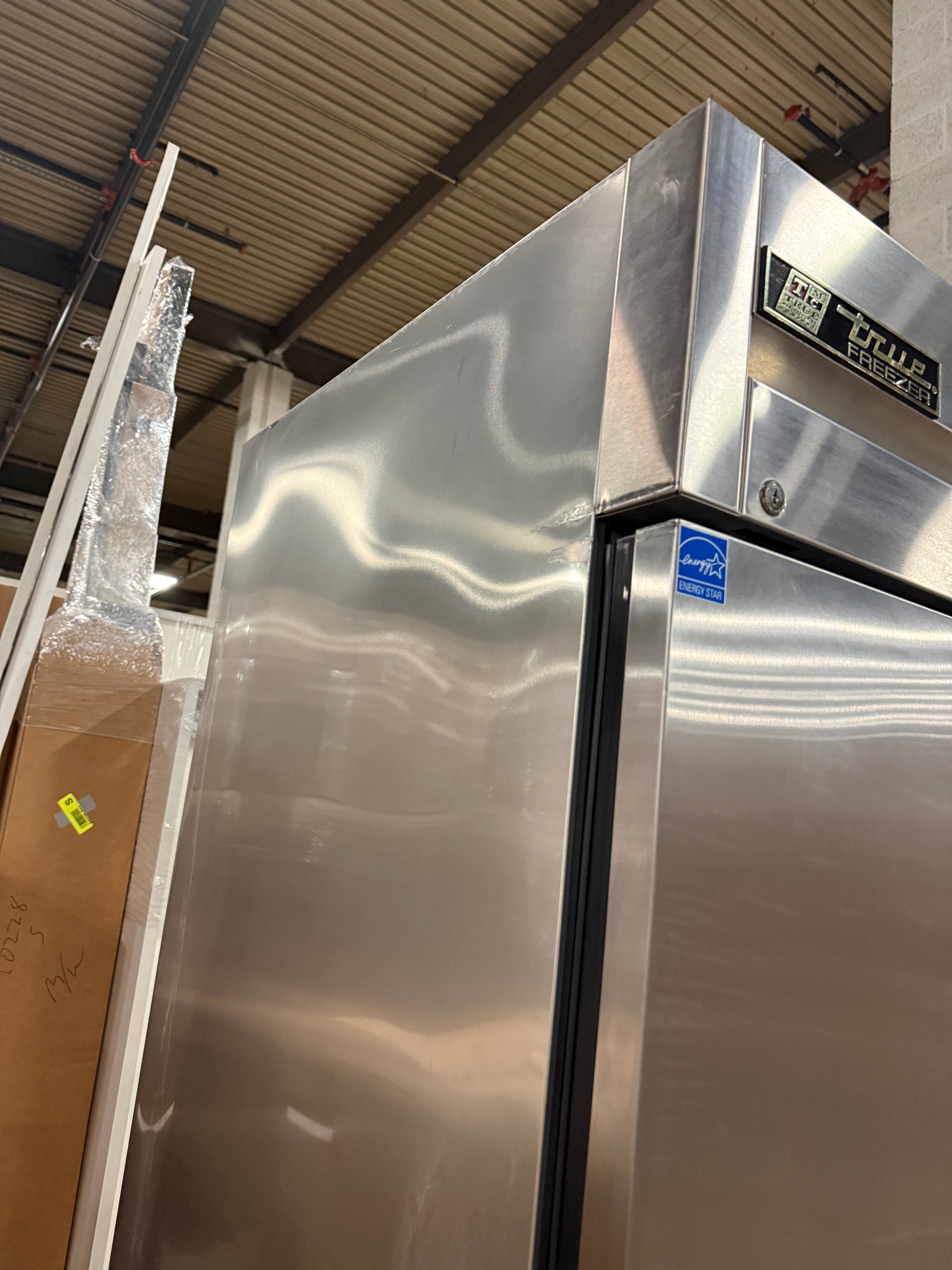 A large stainless steel commercial refrigerator with a black door handle and a visible 'HEALE' brand badge, photographed in a warehouse or showroom setting.
