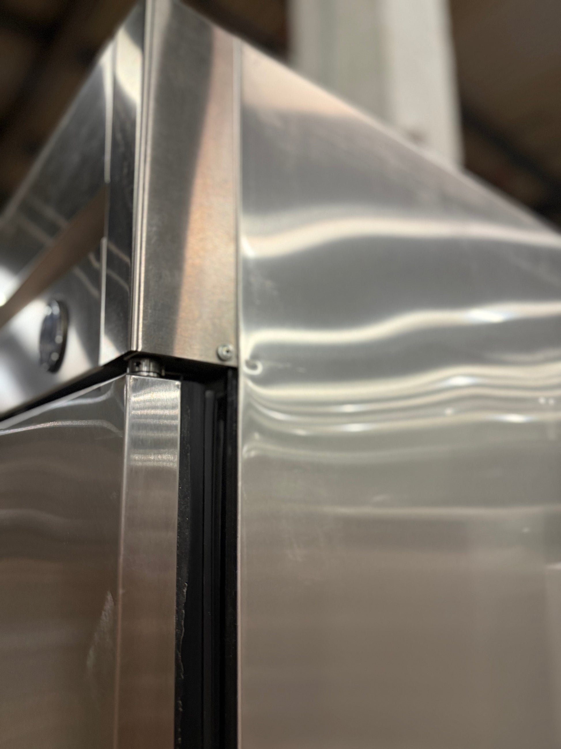 Close-up of a stainless steel appliance, showing the brushed metal surface, corner seam, and a black rubber gasket.