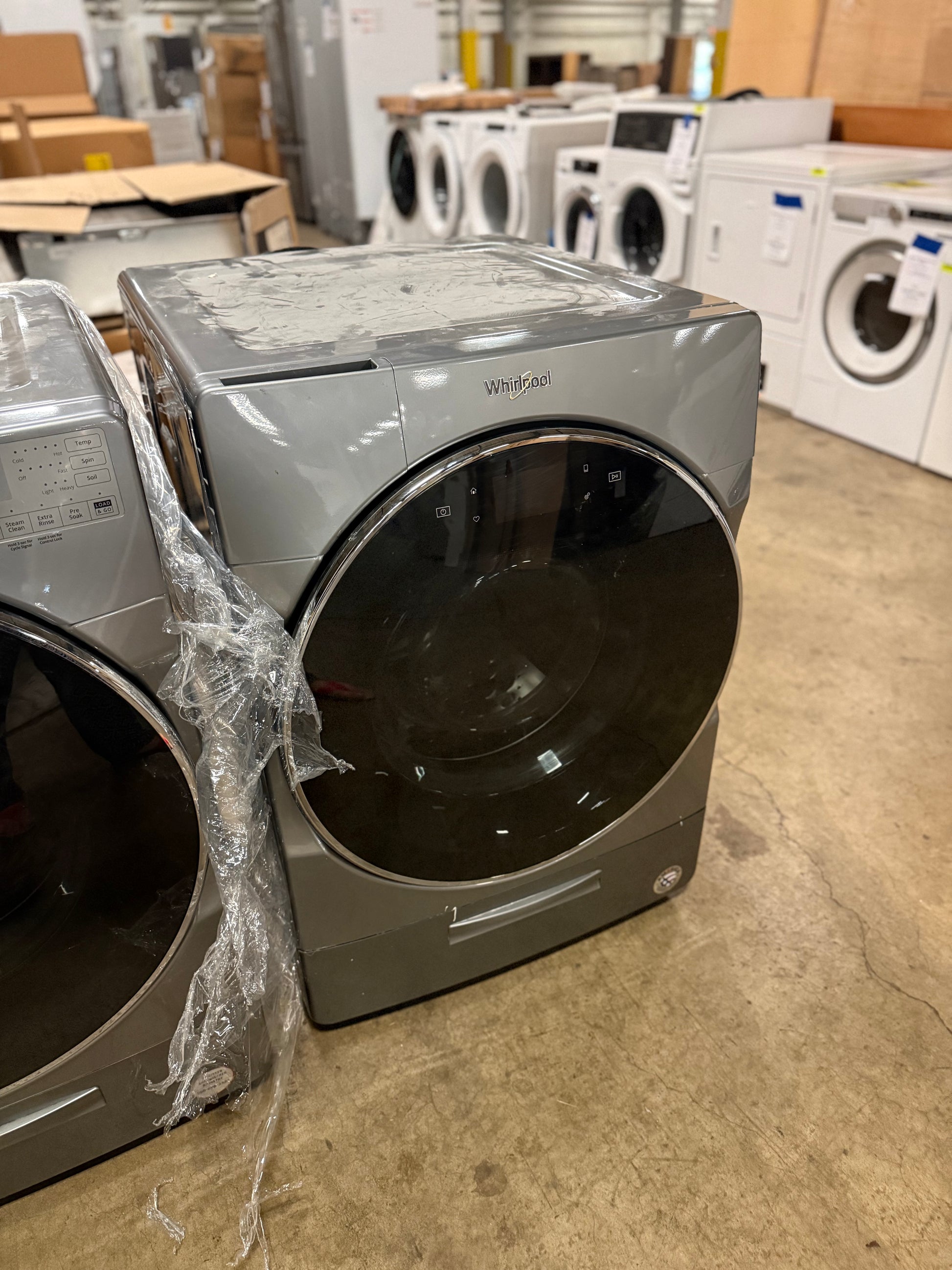 A Whirlpool front-load washer and dryer set in a warehouse or appliance showroom. The dryer is gray with a dark glass door, and the washer is partially visible on the left with plastic wrapping still attached.