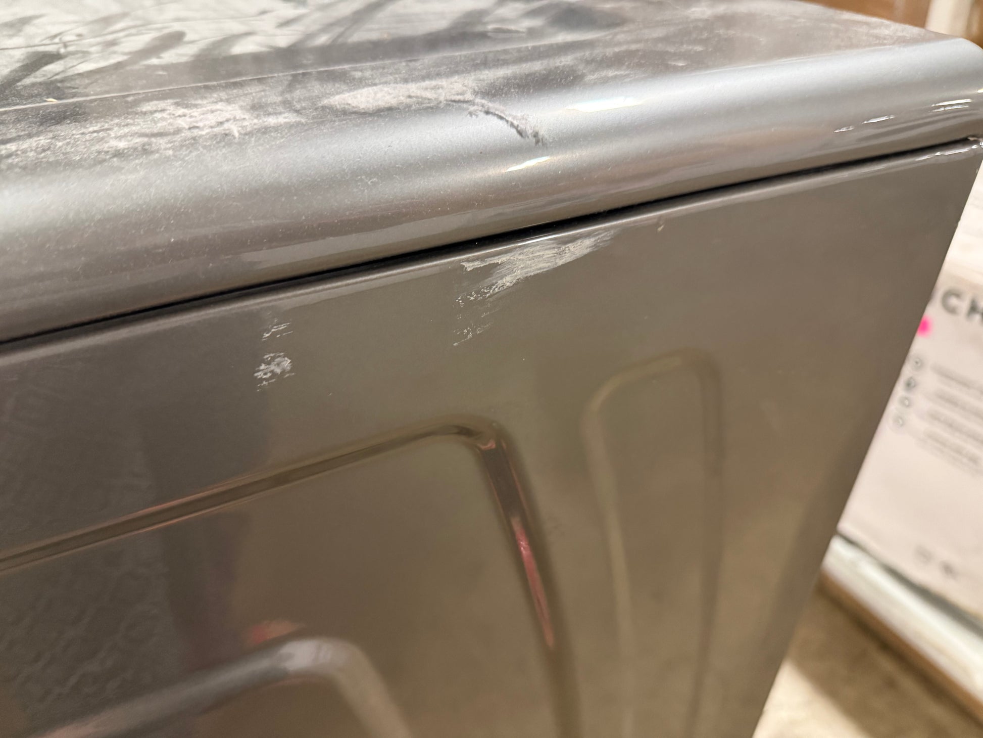 Close-up of the corner of a gray appliance, likely a dryer or washing machine, showing a scuffed metallic surface and seam.