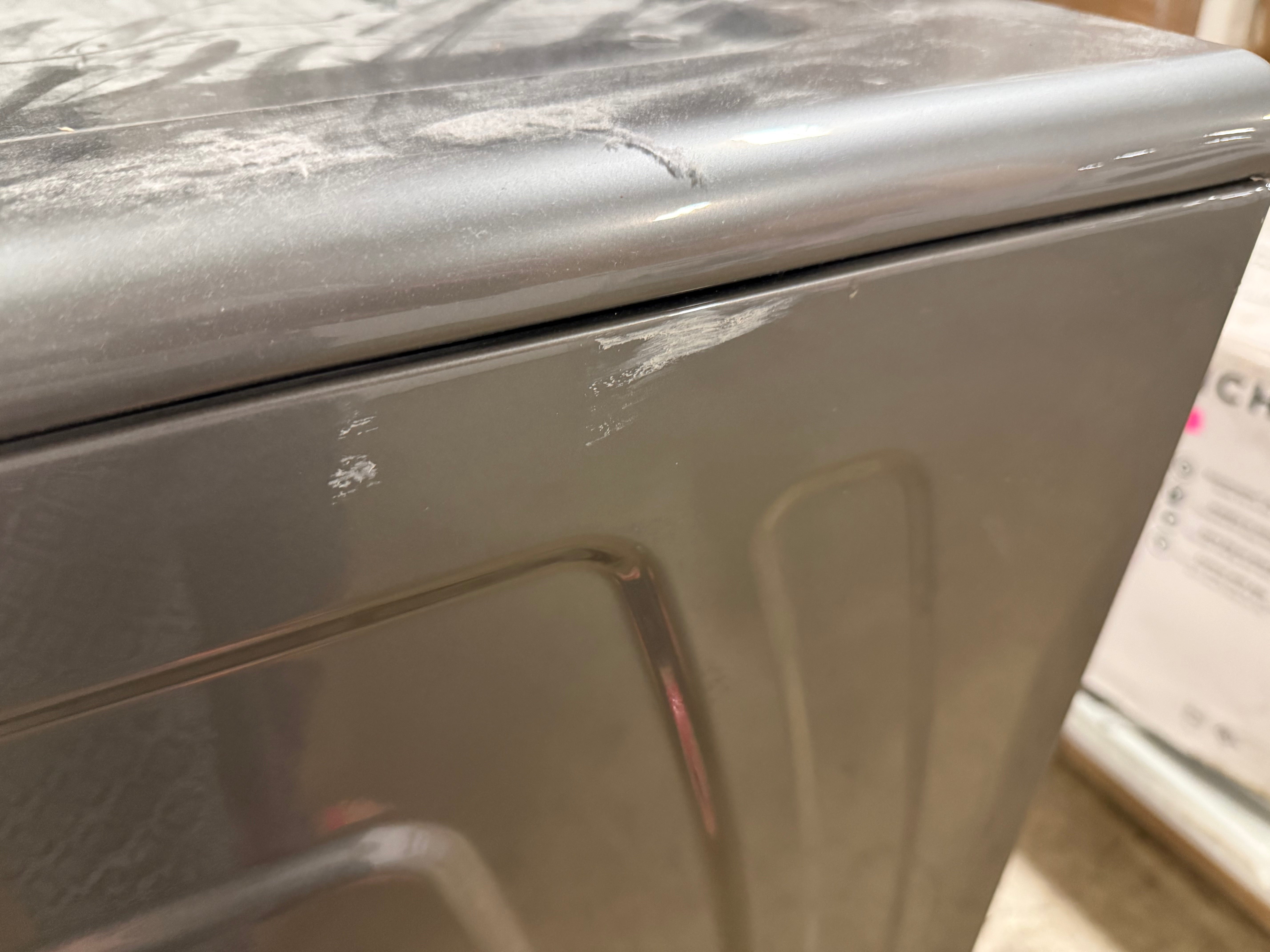 Close-up of the corner of a gray appliance, likely a dryer or washing machine, showing a scuffed metallic surface and seam.