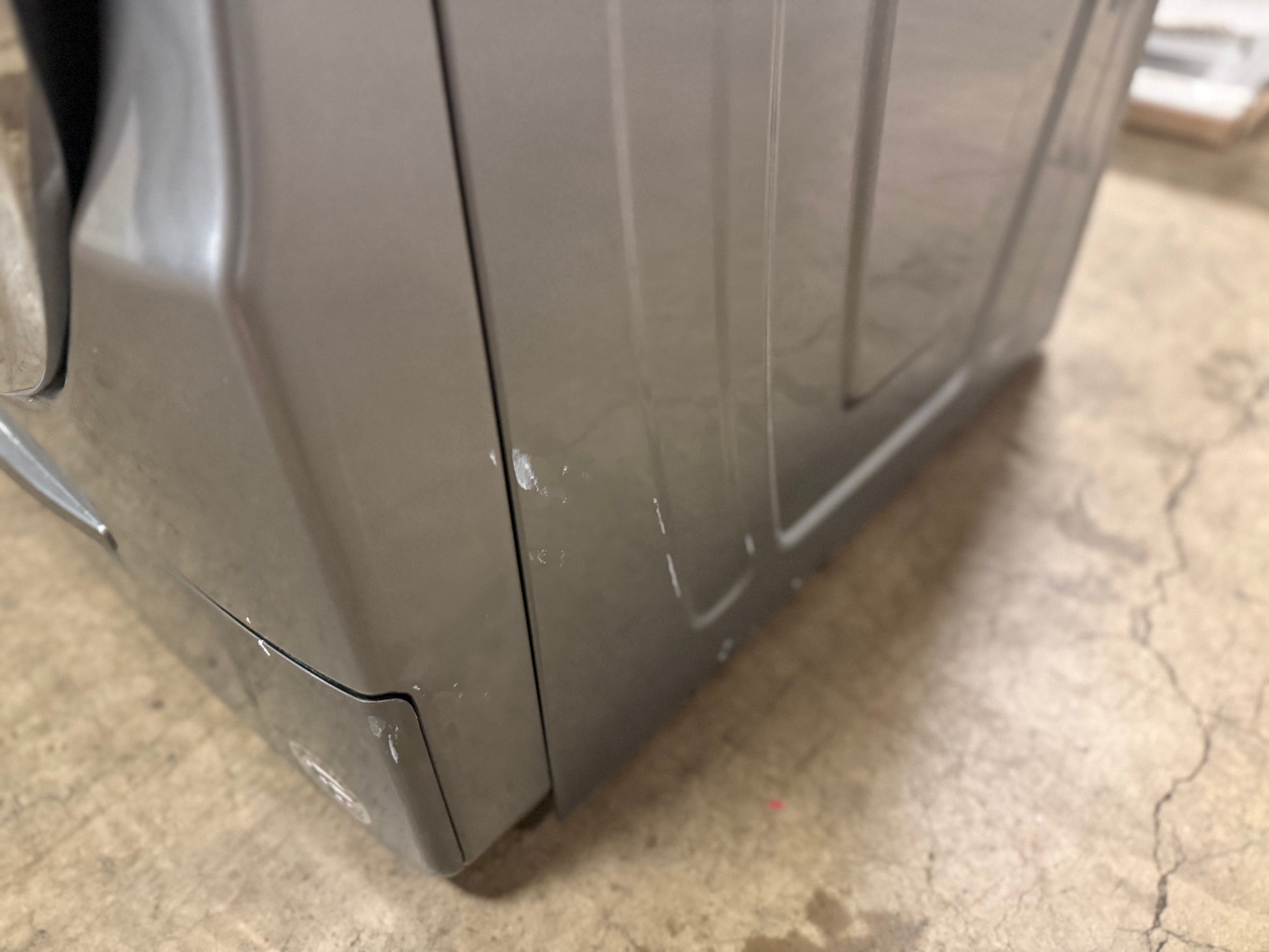 A close-up view of the corner of a gray appliance, likely a washing machine or dryer, showing the side panel and bottom hinge area on a concrete floor.