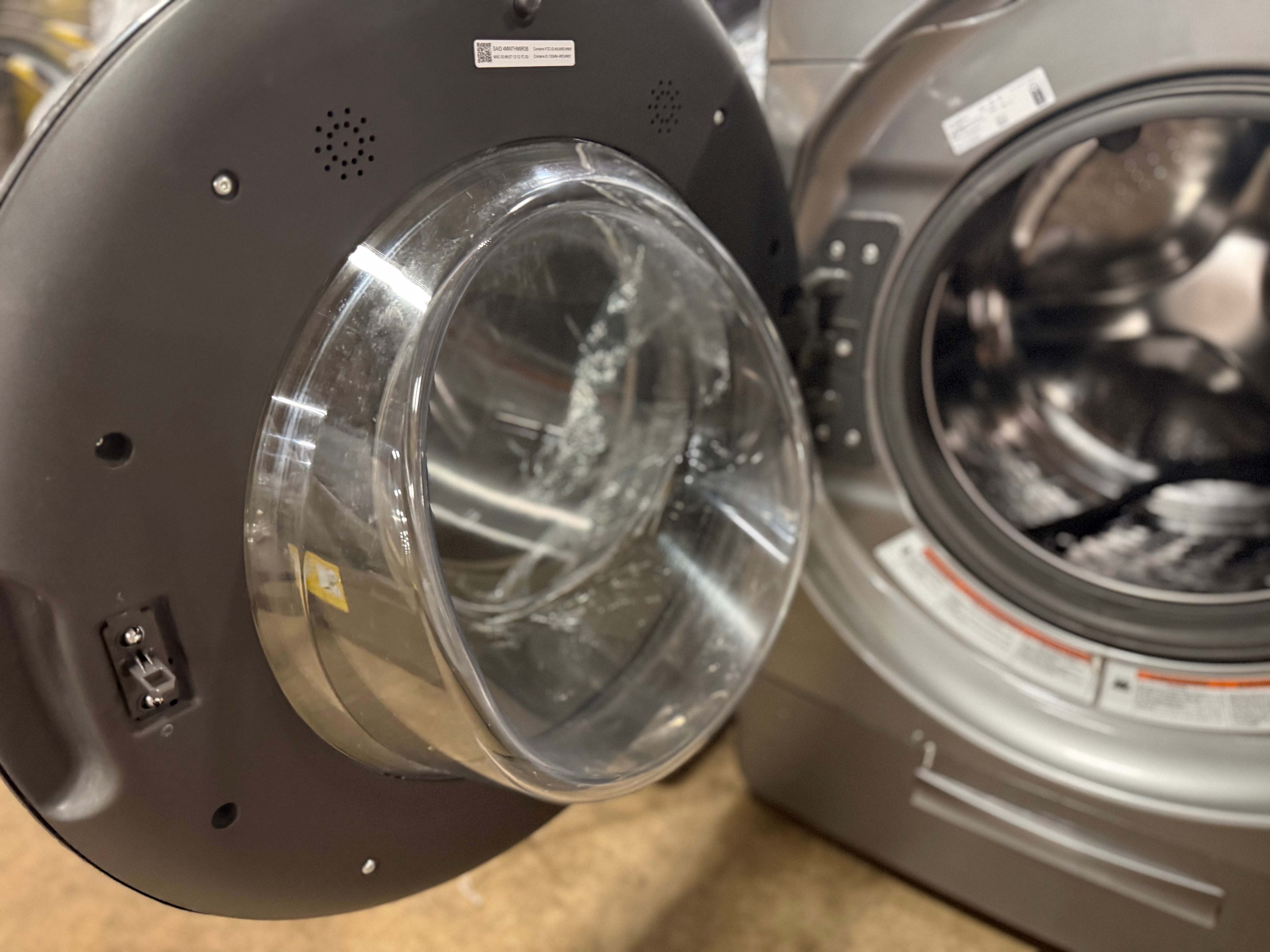 Close-up of a front-loading washing machine with the door open, showing the dark gray exterior, clear circular door seal, and stainless steel drum. Another washing machine is partially visible in the background.