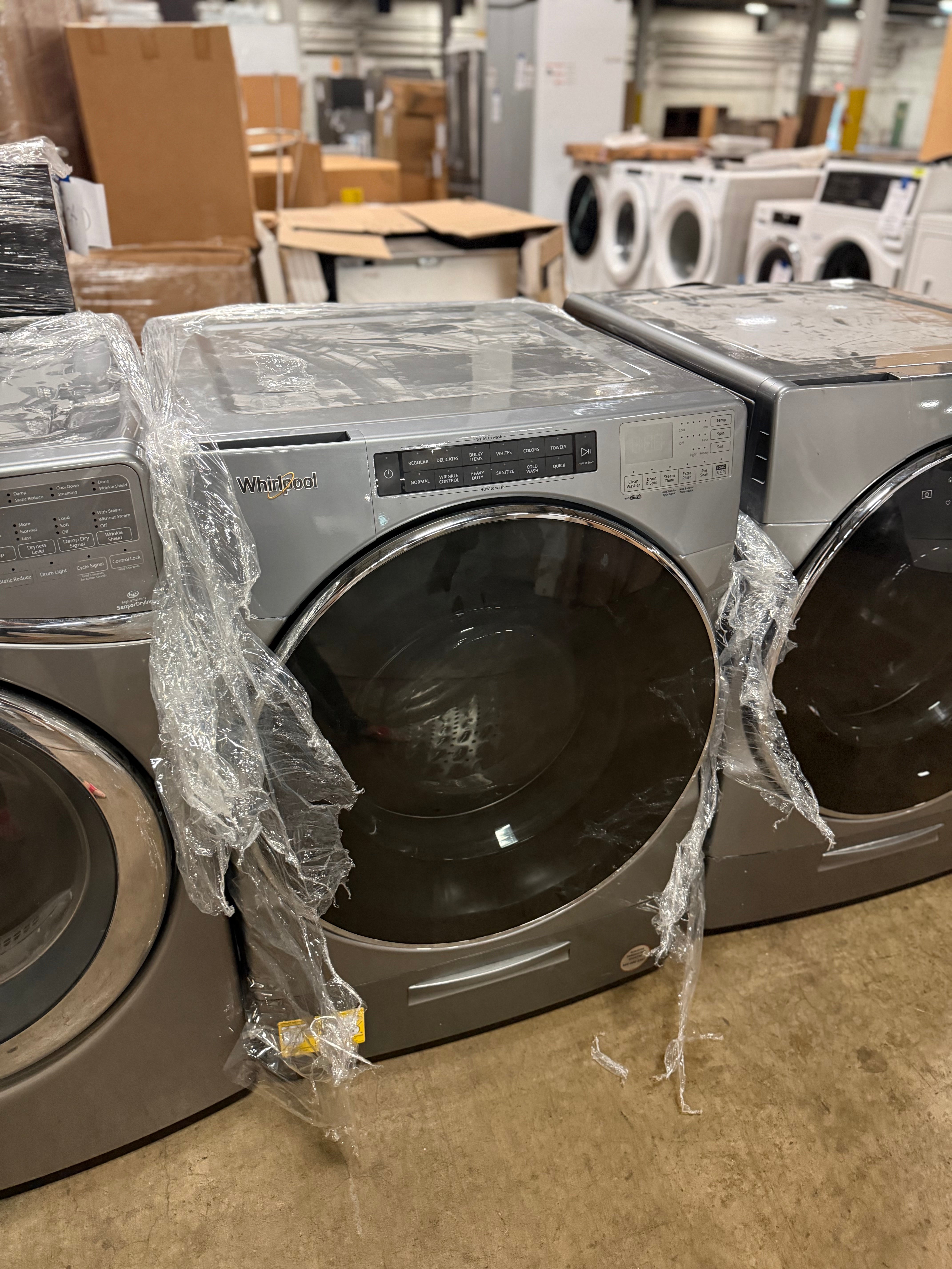 A front-load clothes dryer in a warehouse or appliance showroom setting, wrapped in protective plastic on the sides. The dryer has a metallic gray/silver finish and a large dark glass door.