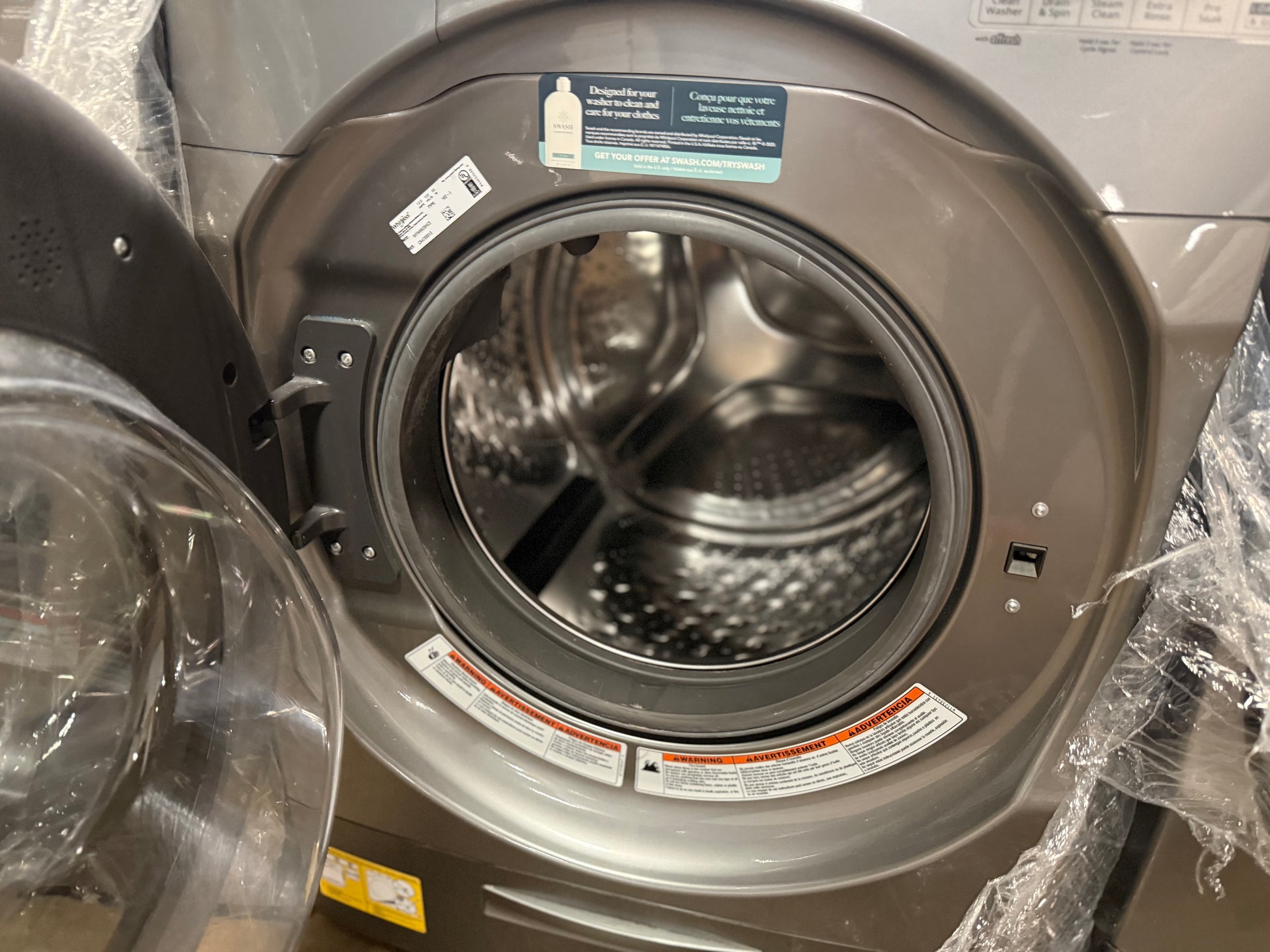 A front-loading washing machine with its door open, showing the stainless steel drum inside. The exterior has a dark gray metallic finish, and protective plastic wrapping is visible around the door and edges.