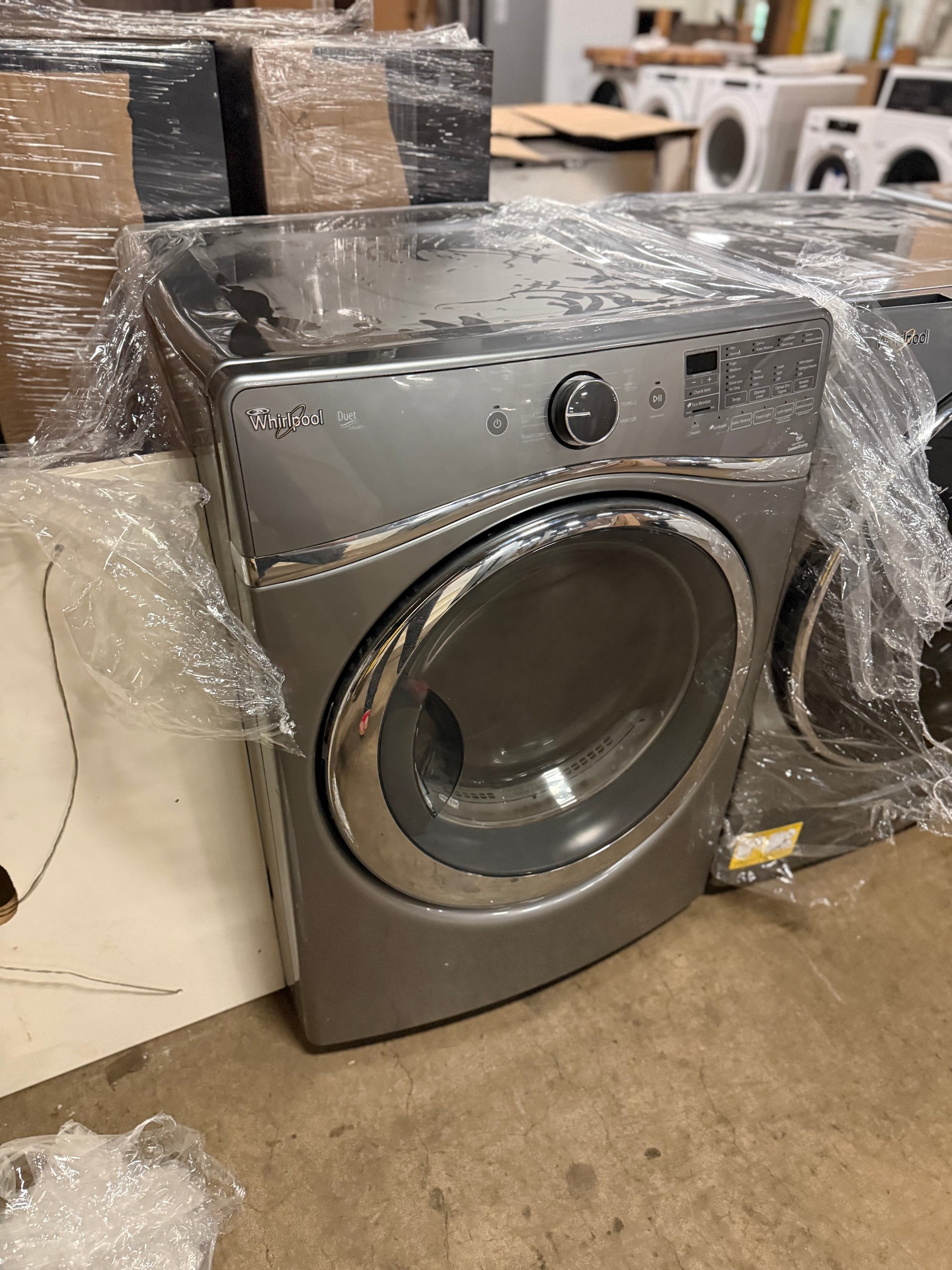 A front-loading Whirlpool washing machine in a brushed stainless steel finish, wrapped in clear plastic film, set in what appears to be a warehouse or appliance showroom.