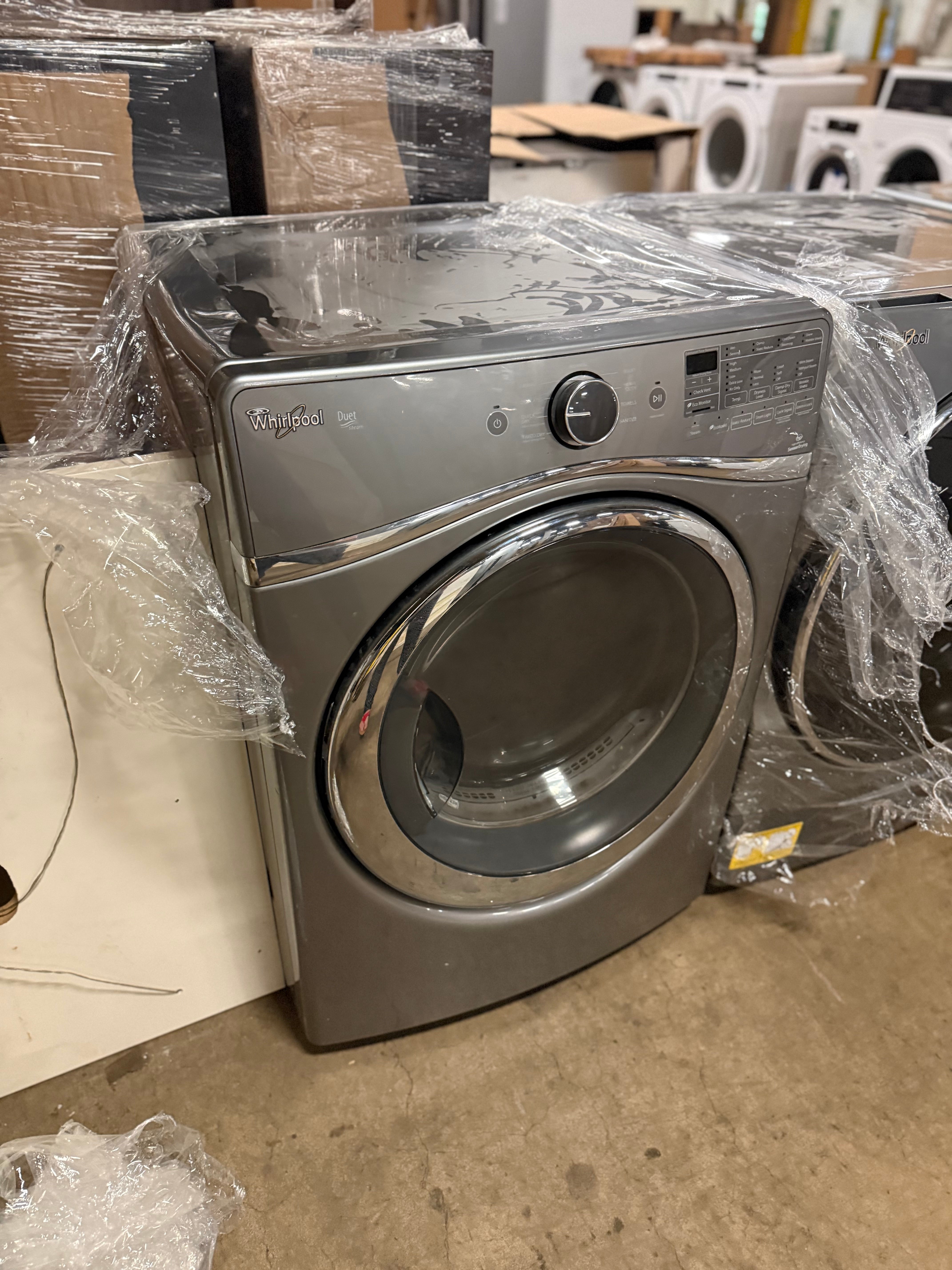 A front-loading Whirlpool washing machine in a brushed stainless steel finish, wrapped in clear plastic film, set in what appears to be a warehouse or appliance showroom.