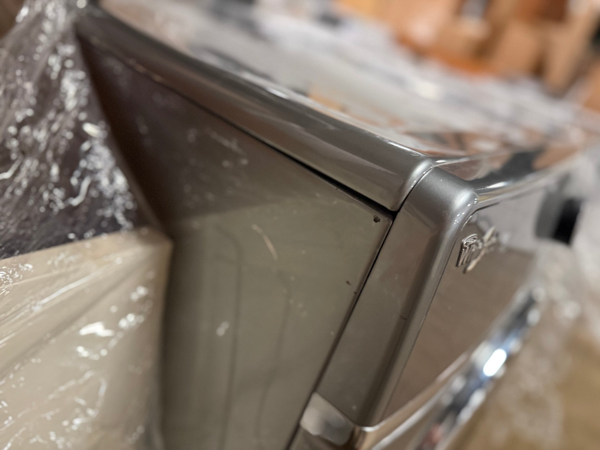 Close-up of a stainless steel appliance corner, likely a coffee machine or espresso machine, showing the side panel and top edge with some protective plastic wrapping visible.