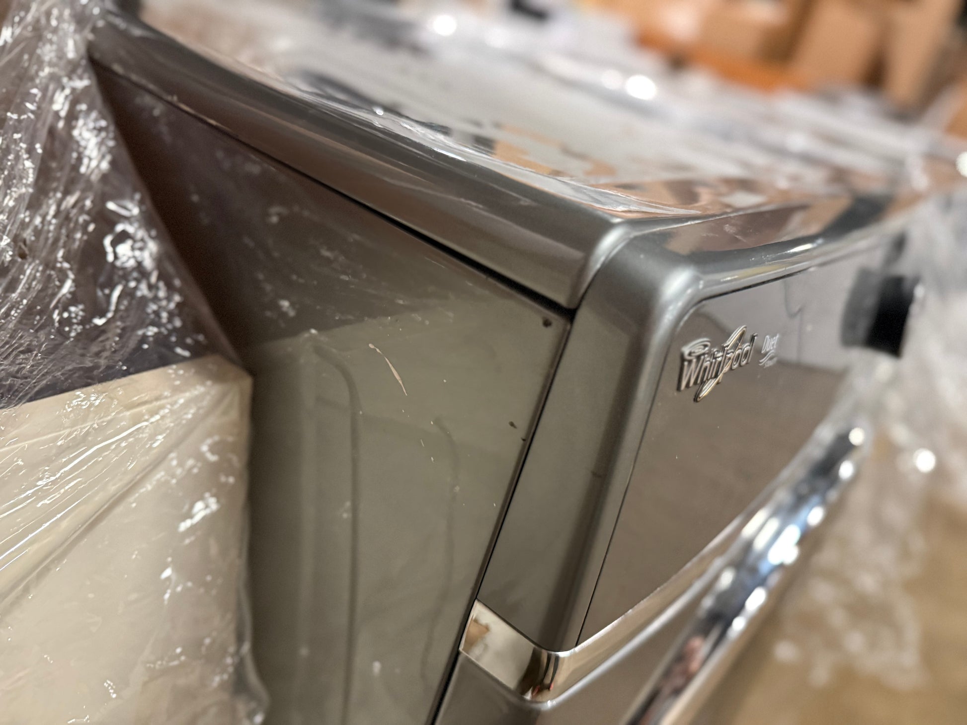 A close-up of a Whirlpool appliance, likely a refrigerator or freezer, wrapped in protective plastic film. The metallic gray side panel features the silver Whirlpool logo, and a chrome handle is partially visible in the foreground.