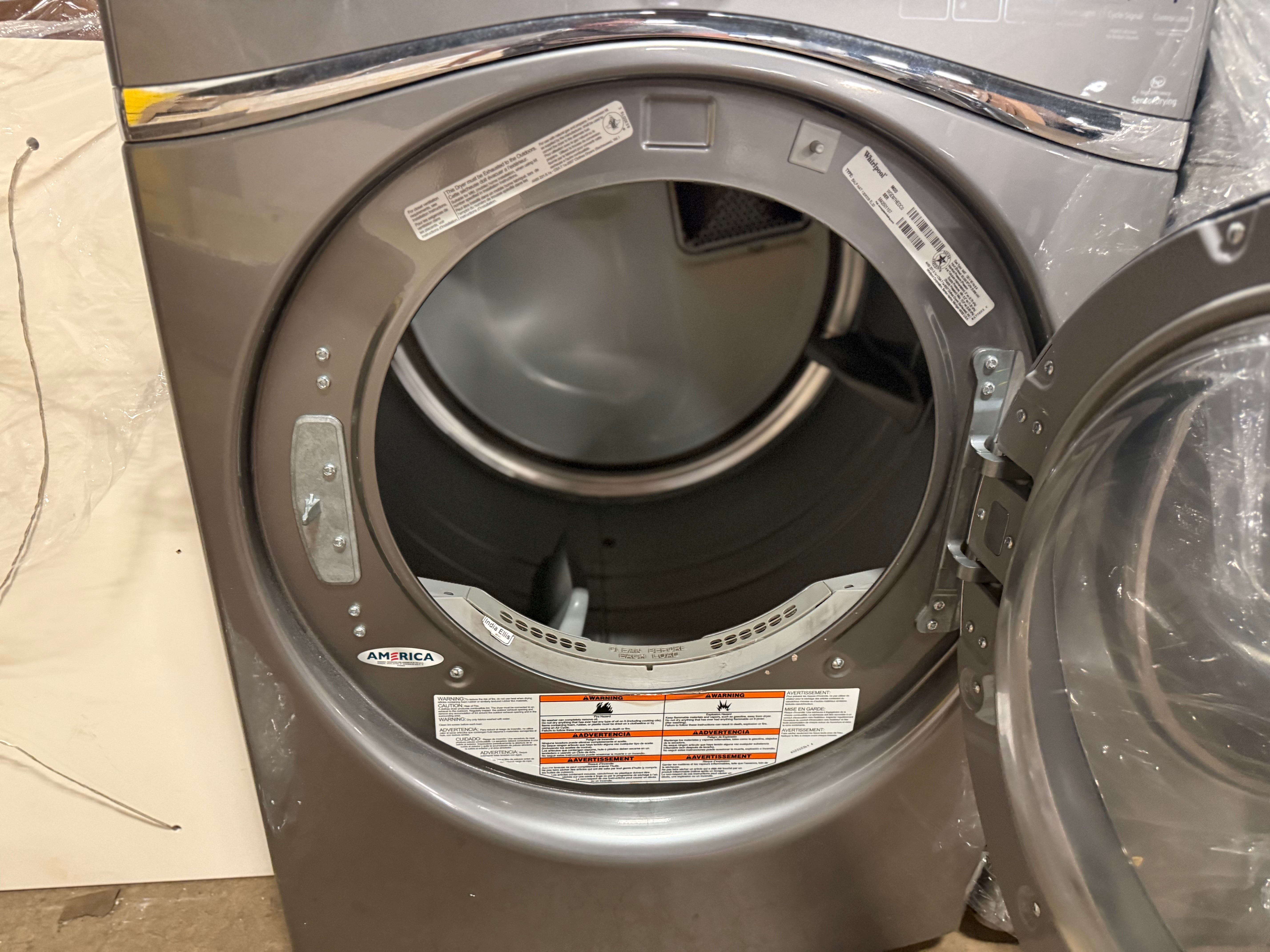 A front-loading clothes dryer with its door open, showing the interior drum and control panel. The appliance has a metallic gray finish and is photographed in a warehouse or showroom setting.