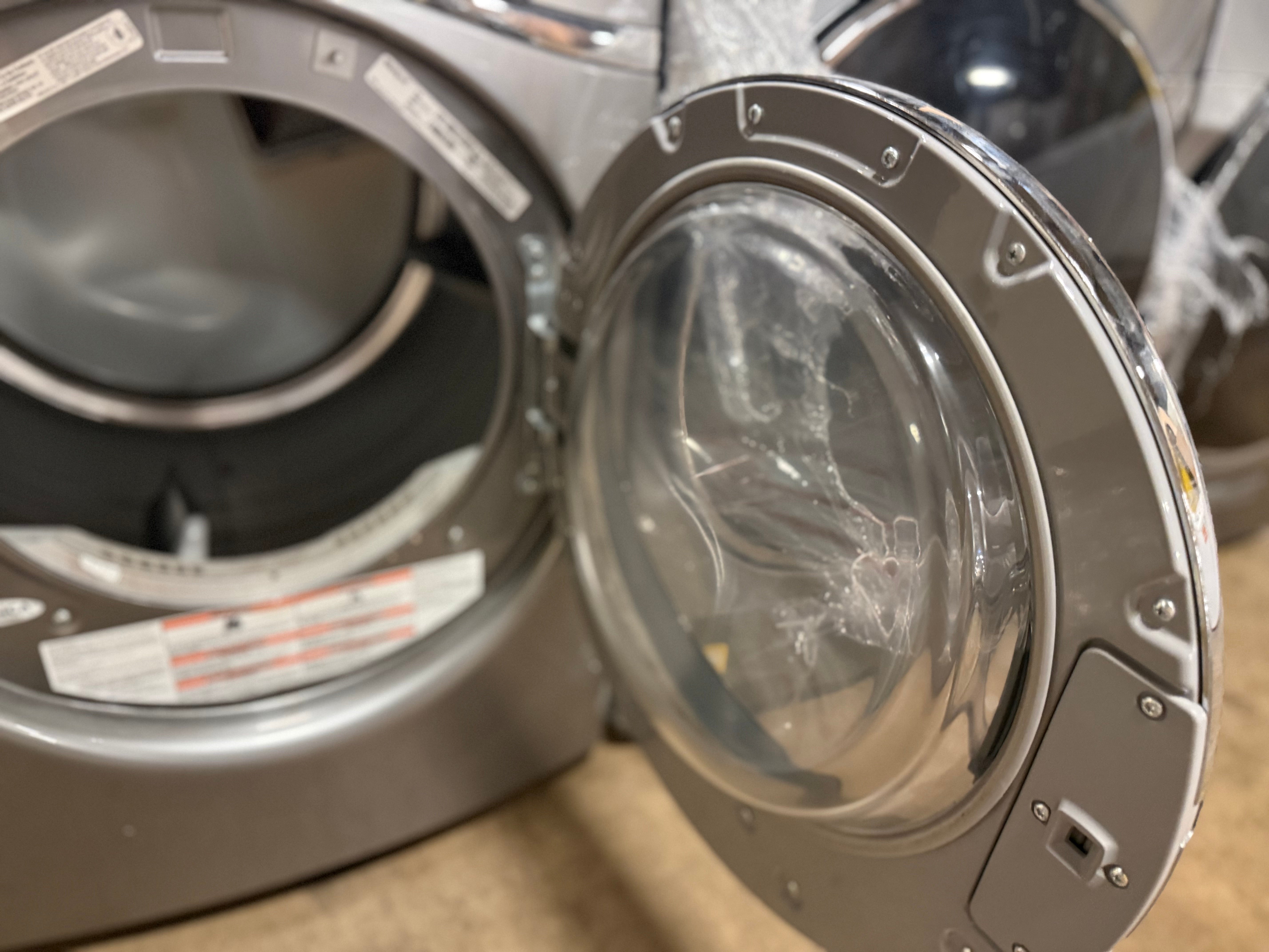 A close-up of a front-loading washing machine with the door open, showing the silver exterior, clear door, and inner drum.
