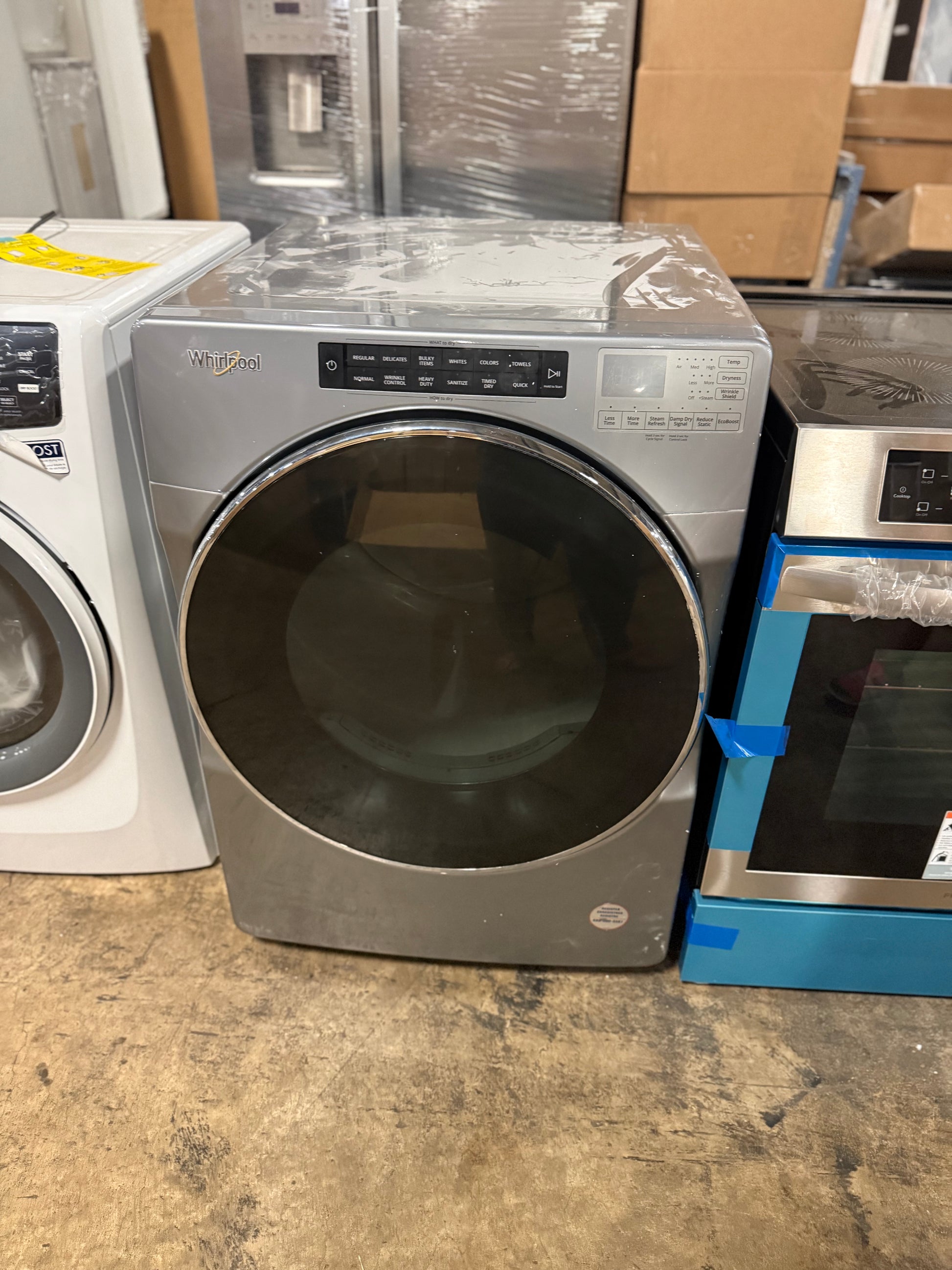A front-load washing machine in a warehouse-like setting, with a large dark glass door and a control panel on top. A white washing machine is partially visible on the left, and a blue appliance is partially visible on the right.