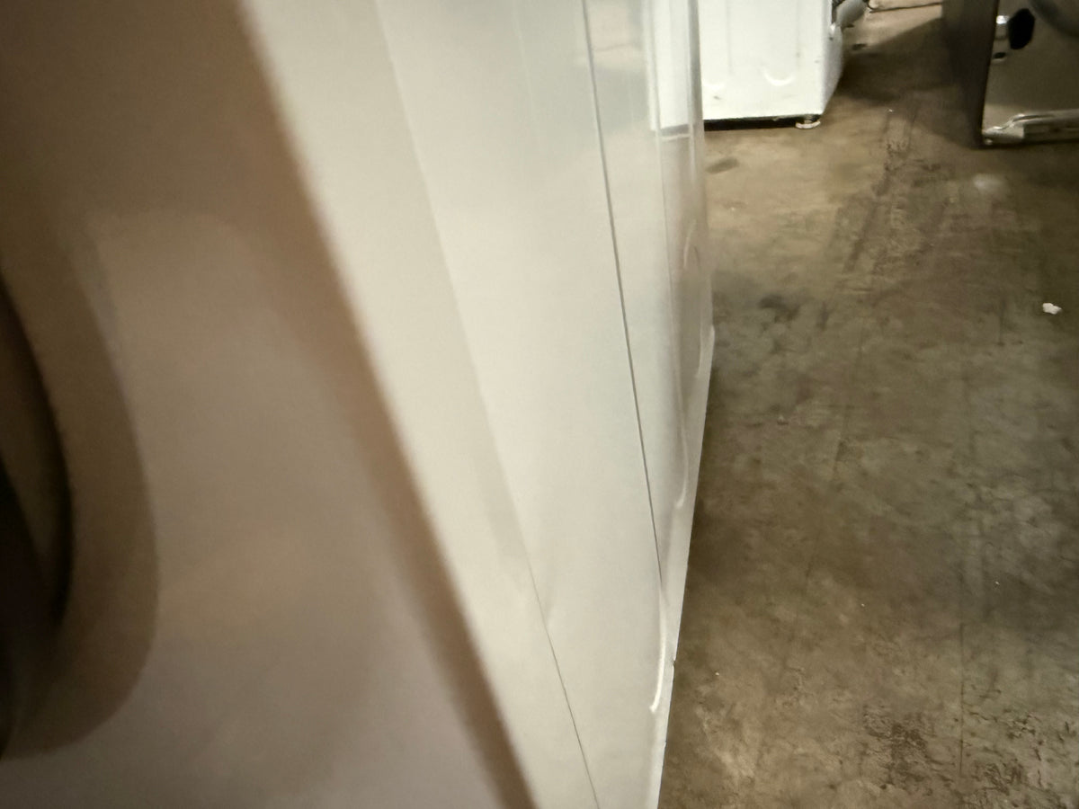 A close-up view of a white appliance, likely a washing machine or dryer, showing its side panel and door edge, with a concrete floor and other appliances in the background.