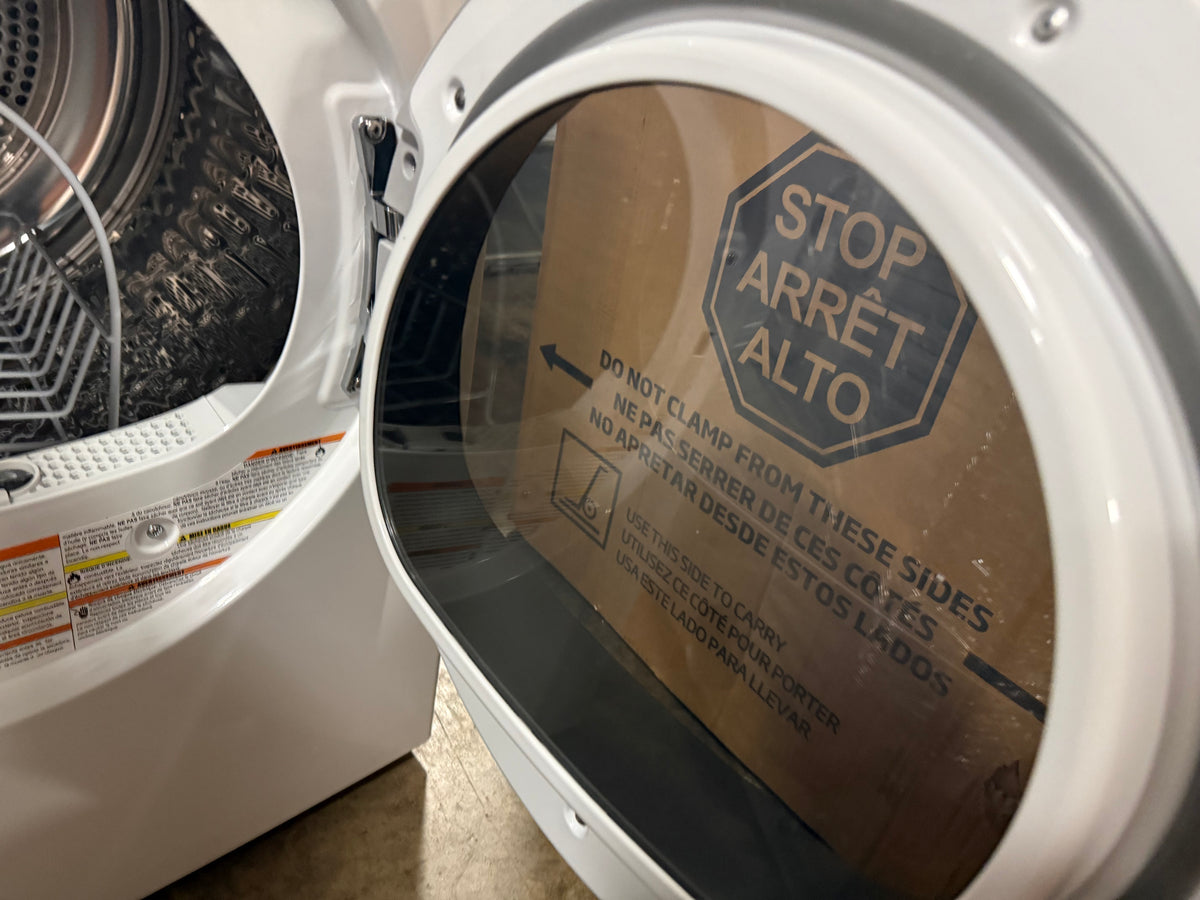 Close-up of a white front-loading washing machine door with the glass door open, showing the drum inside and a cardboard protective sheet covering part of the door with printed warning text.