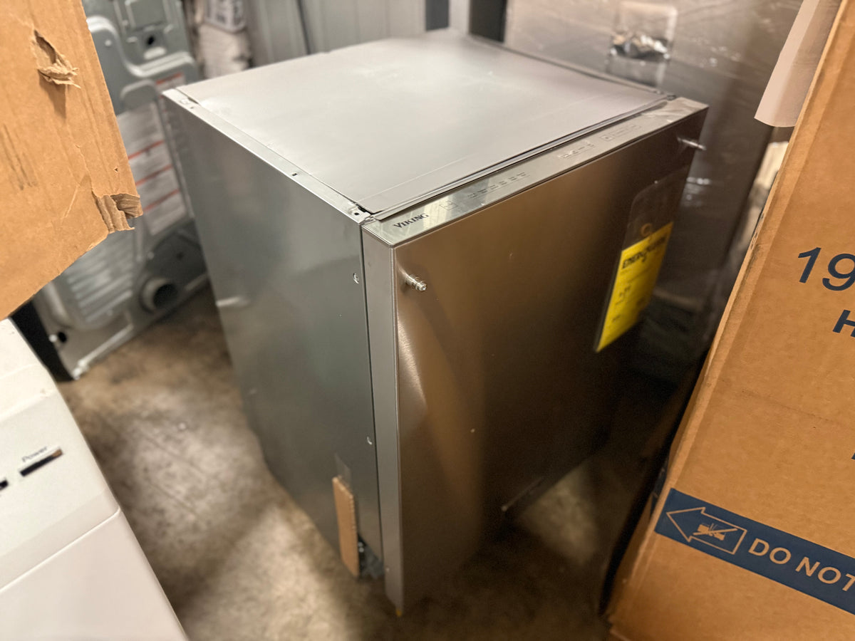 A stainless steel appliance, likely a dryer or similar home appliance, standing upright in a warehouse or storage area surrounded by cardboard boxes.