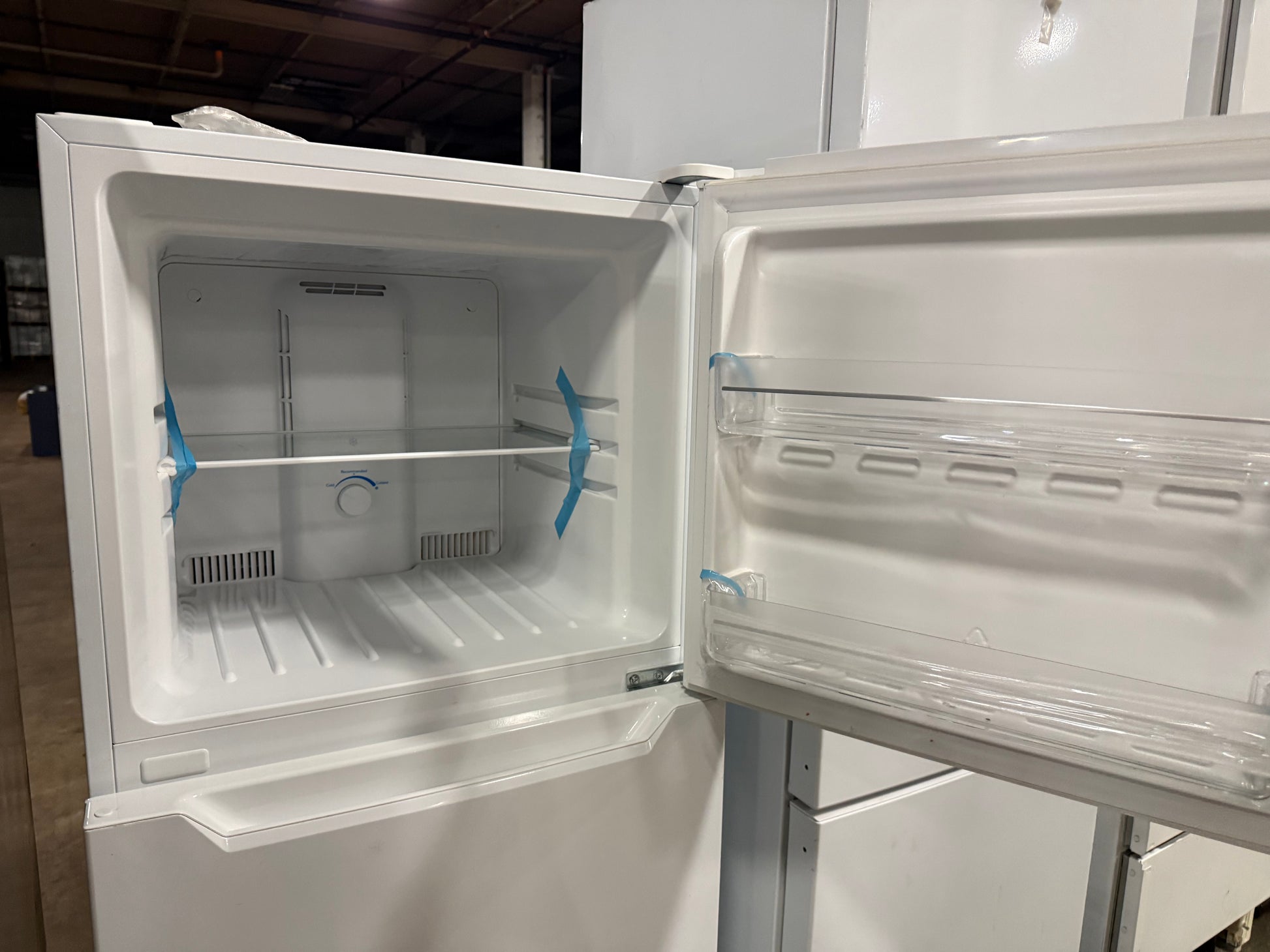 A white top-freezer refrigerator shown with the freezer door open, revealing an empty white interior with clear plastic shelves and blue plastic grommets, photographed in what appears to be a warehouse or showroom.