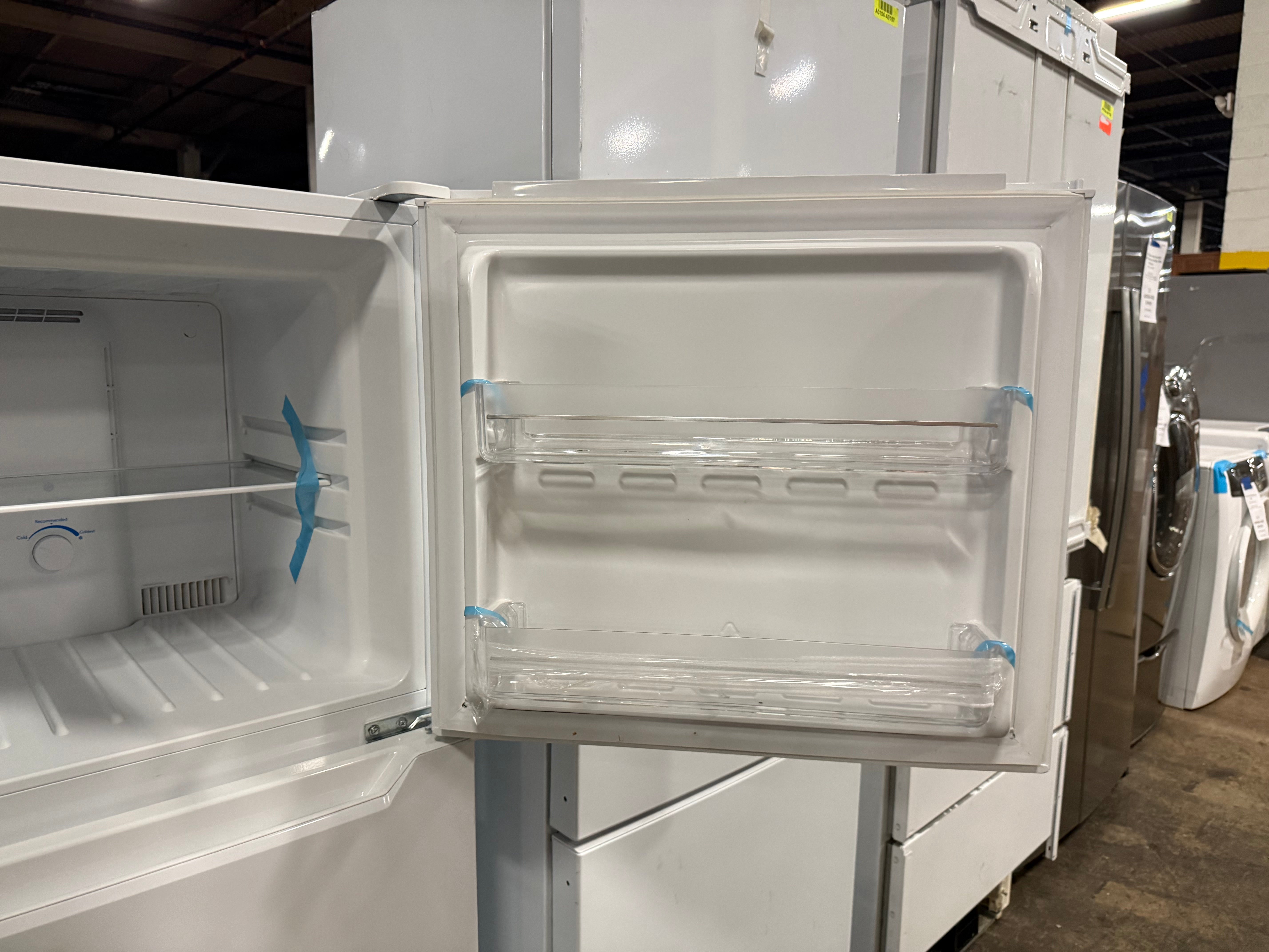 A white chest freezer shown with its door open, revealing a clean interior with two clear glass door bins and a blue plastic fan. Other appliances are visible in the background in what appears to be a showroom or warehouse setting.