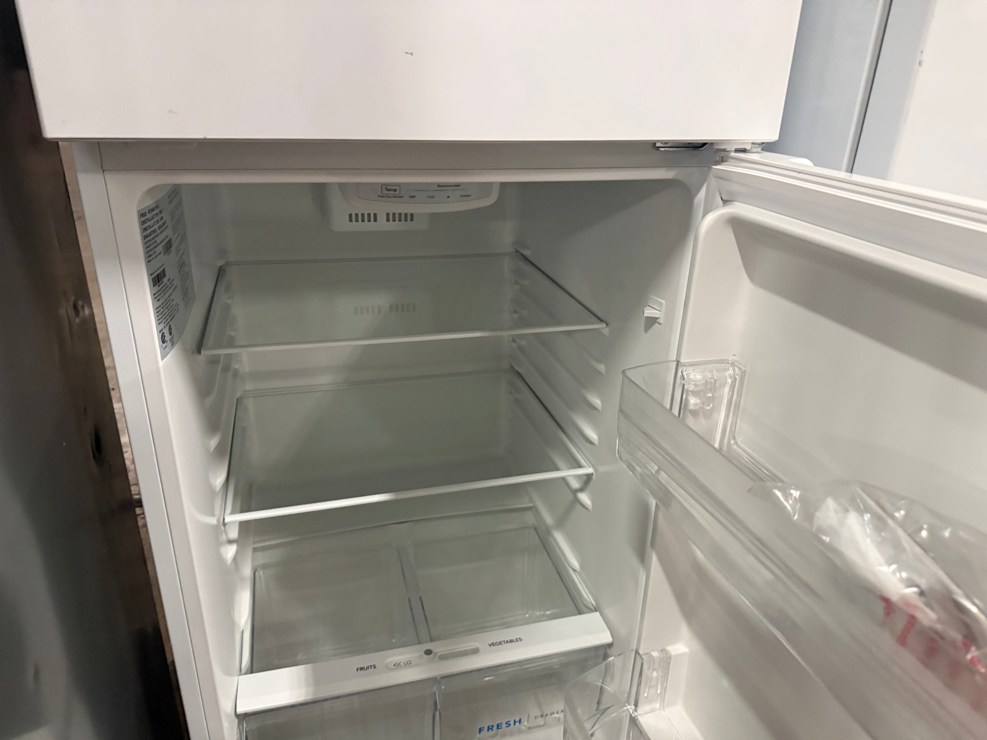 An empty white refrigerator with its door open, showing clear glass shelves, a lower crisper drawer, and a plastic bag of produce on the door shelf.