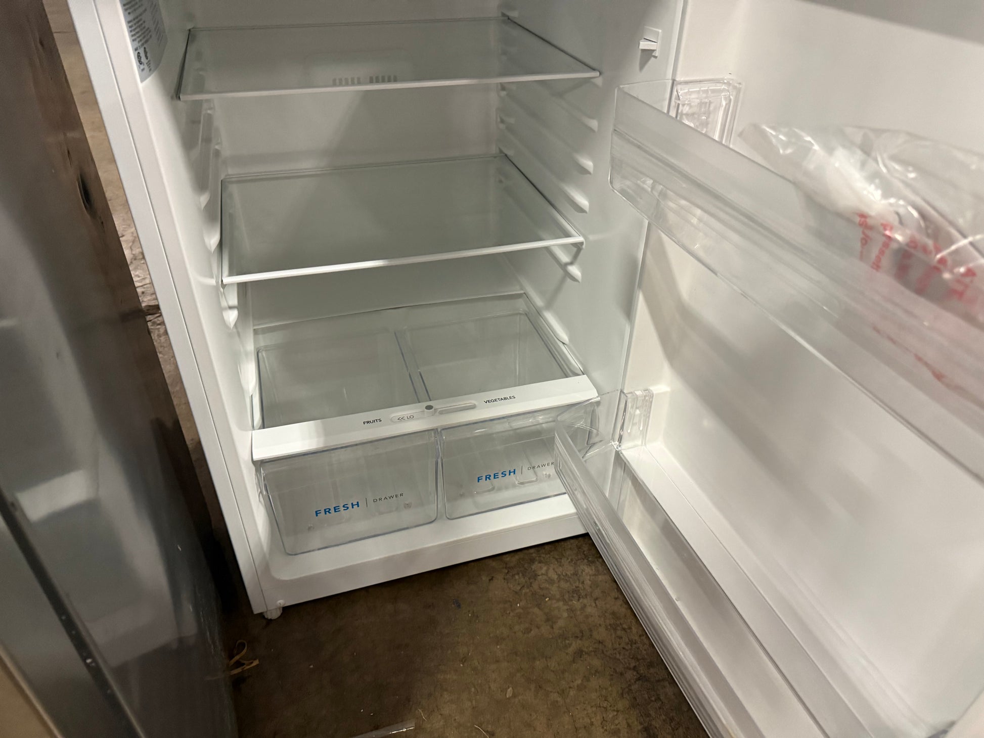An empty white refrigerator with its door open, showing clear glass shelves, two clear plastic drawer bins labeled FRESH, and a plastic bag containing produce on the door shelf.