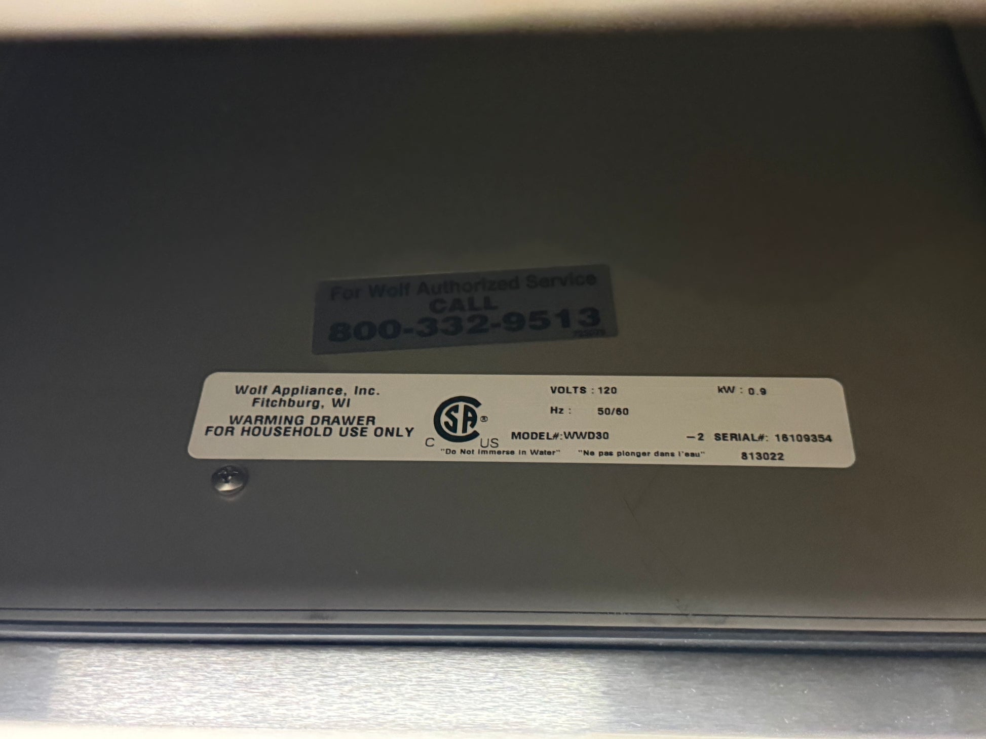 A close-up of a metallic appliance surface with a white product label reading Wolf Appliance, Inc., model WV6030, and a blue service call sticker above it.