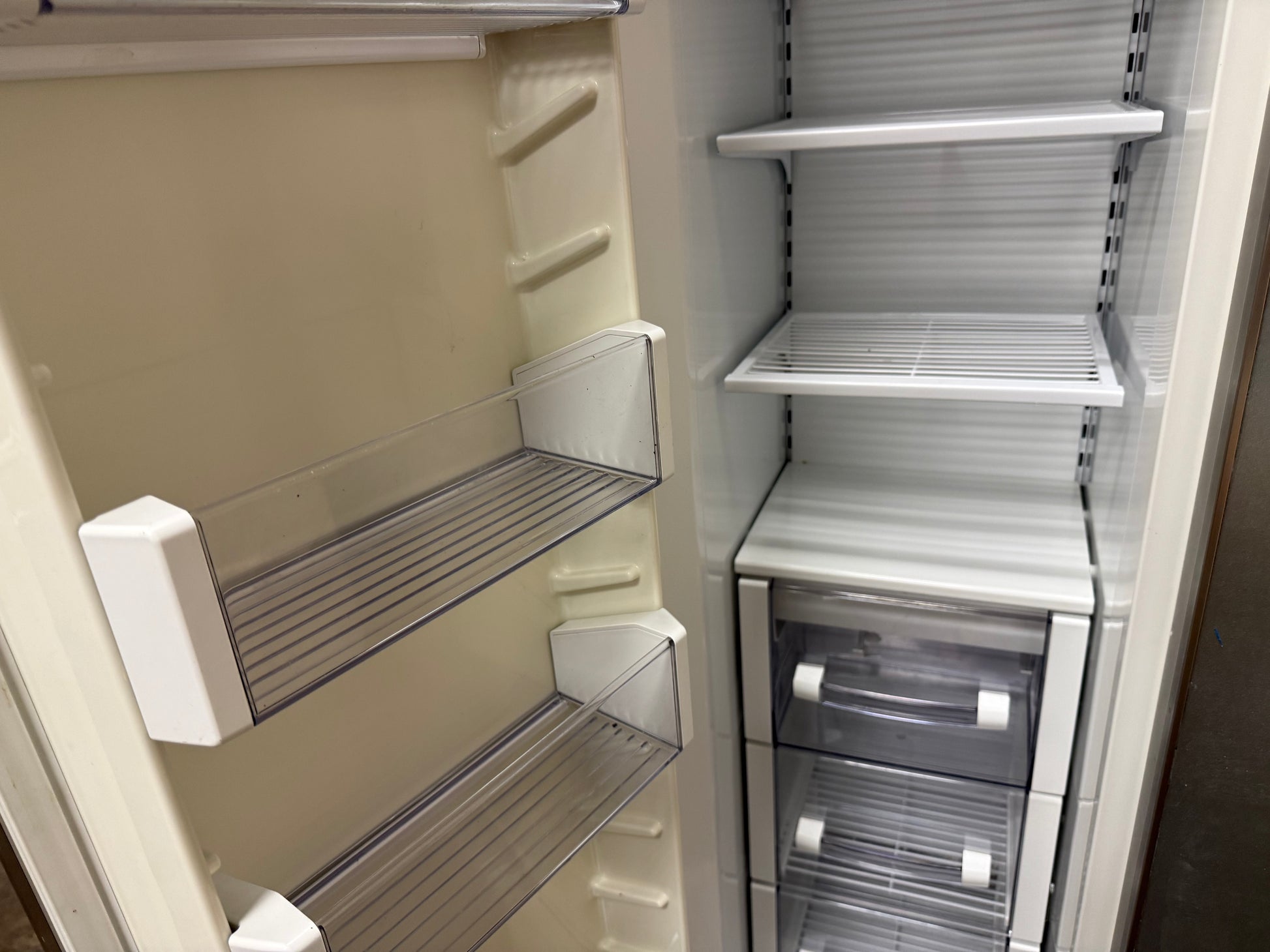 An empty refrigerator interior with clear glass shelves, white door bins, and two lower transparent drawers on a light neutral background.