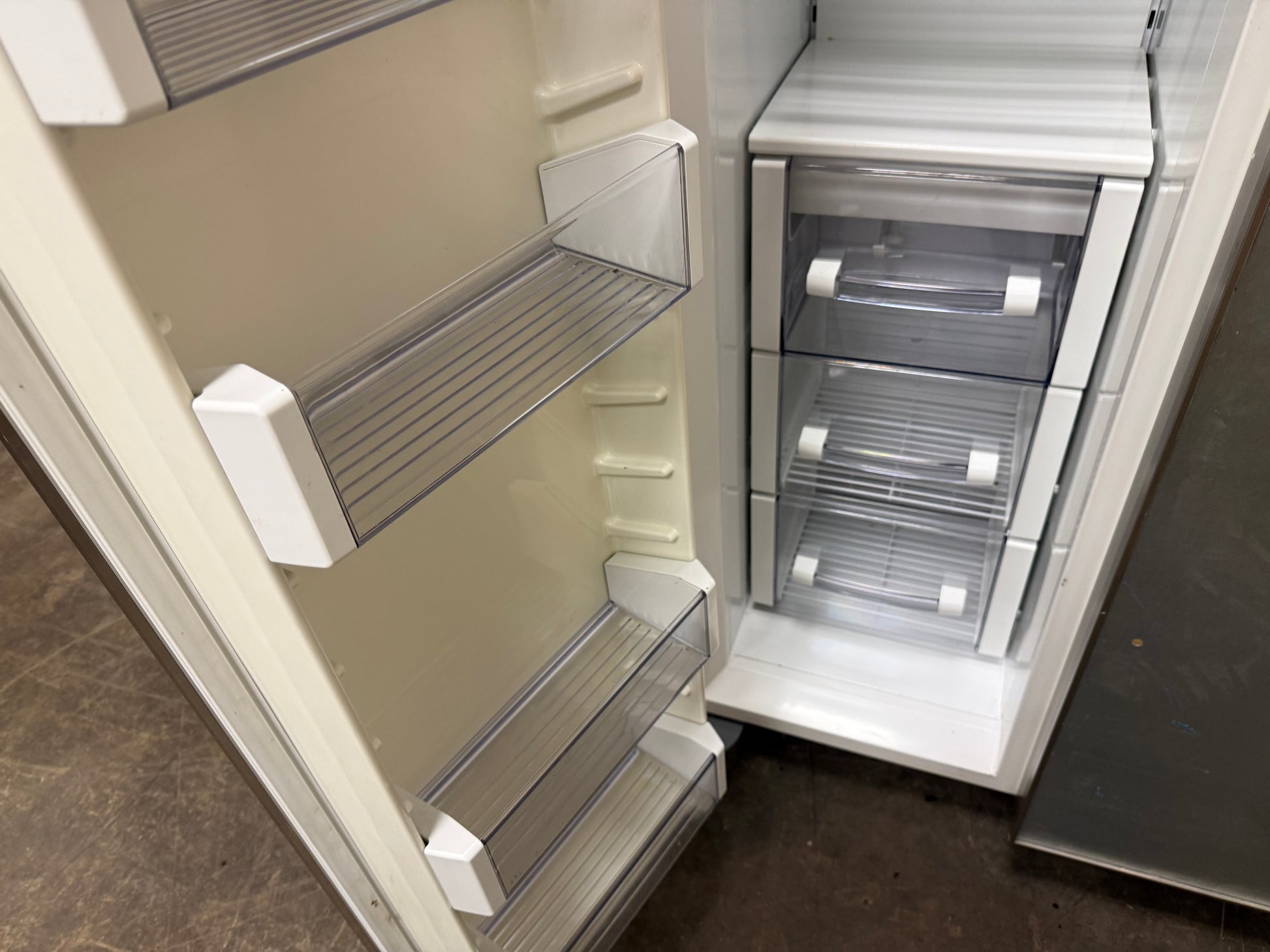An open refrigerator showing the interior with clear glass door bins on the left and three transparent drawer compartments on the right, photographed in a real indoor setting with a dark floor visible.
