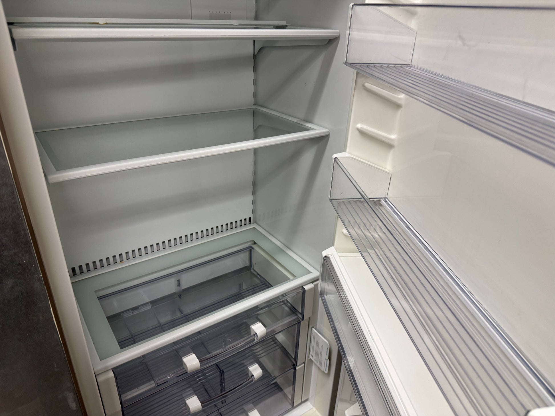 An empty white refrigerator interior with glass shelves, door bins, and a lower crisper drawer compartment.