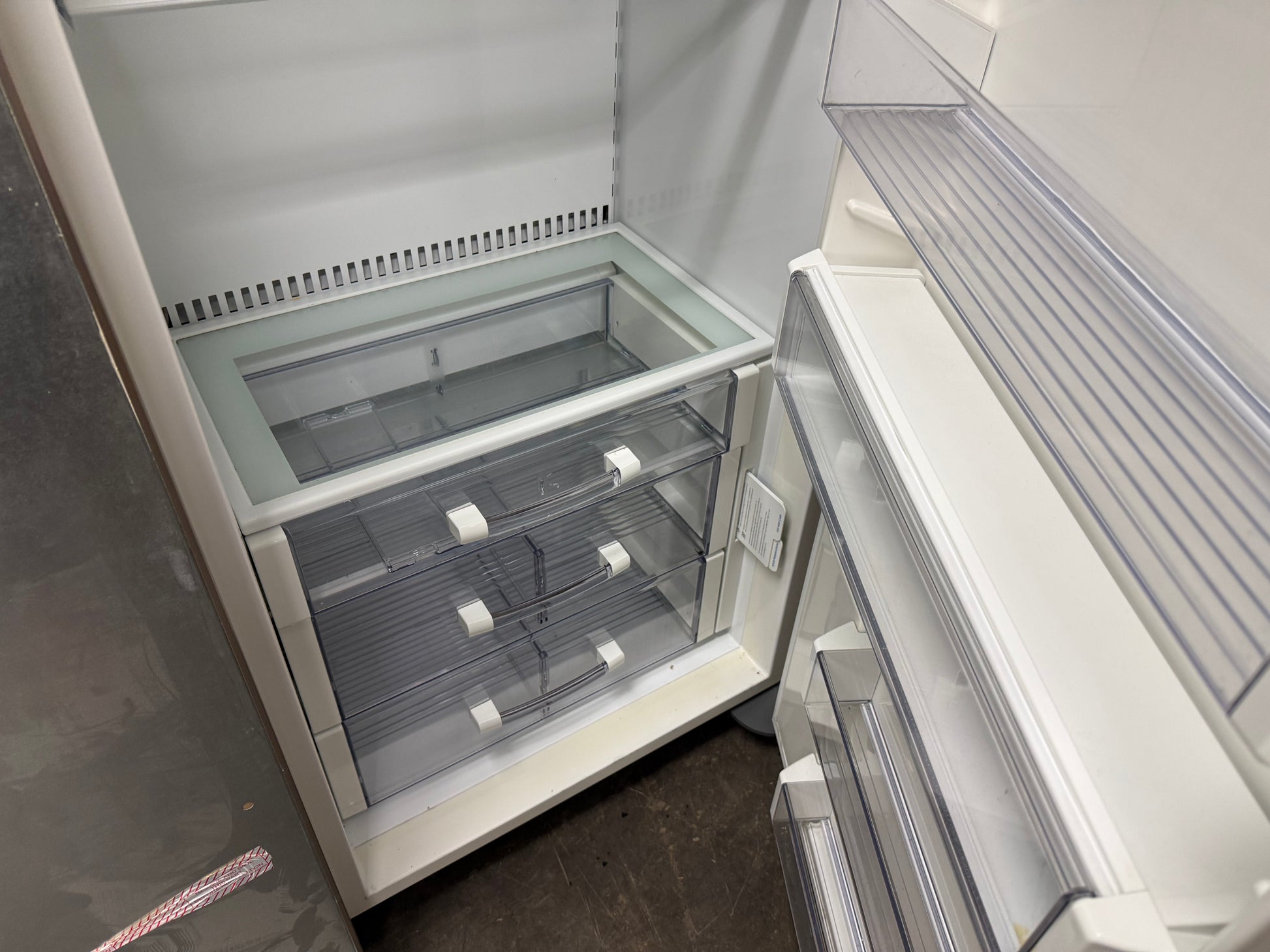 An empty white refrigerator with the lower freezer drawer open, showing clear plastic storage bins and interior shelves.