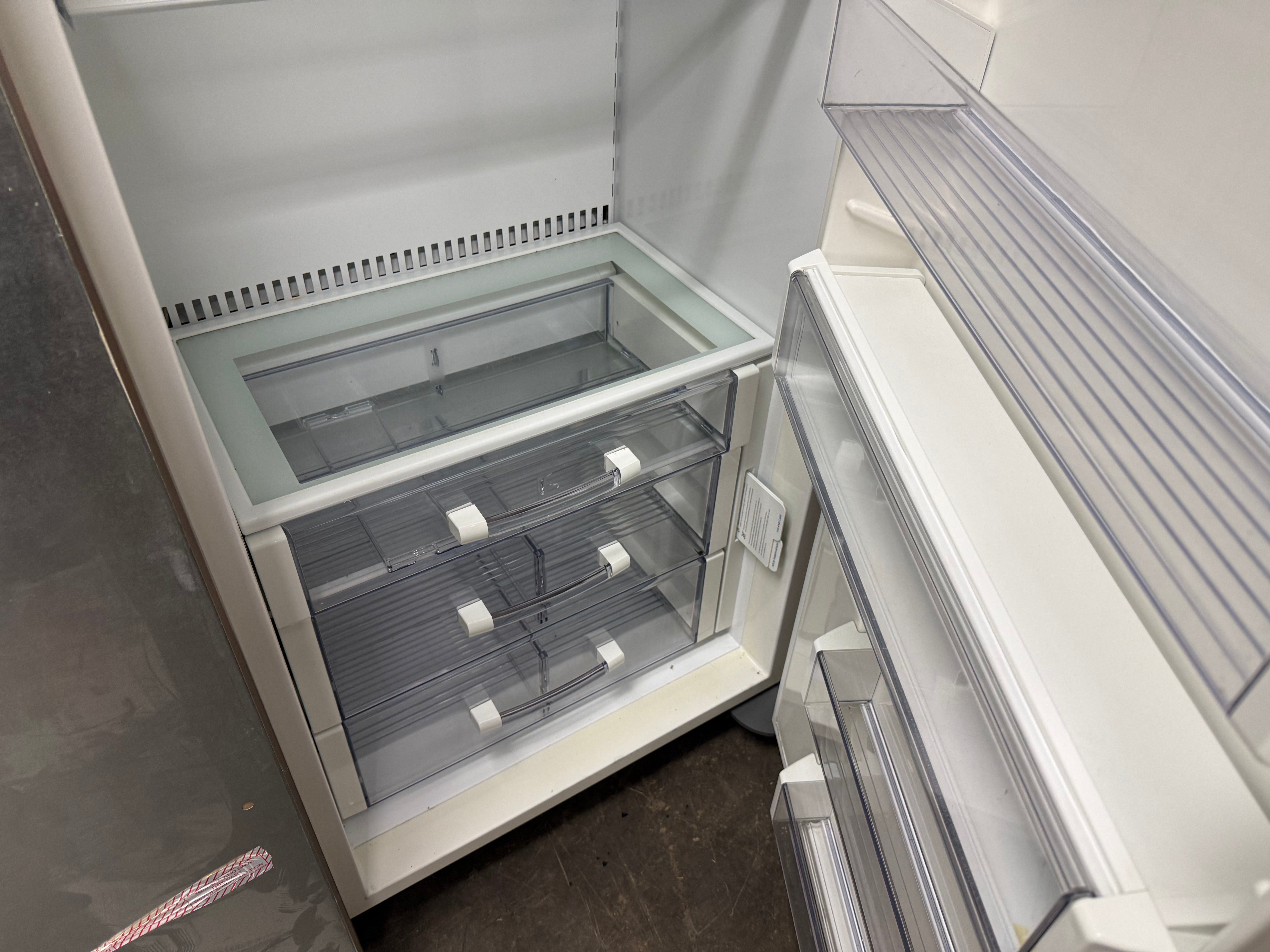 An empty white refrigerator with the lower freezer drawer open, showing clear plastic storage bins and interior shelves.