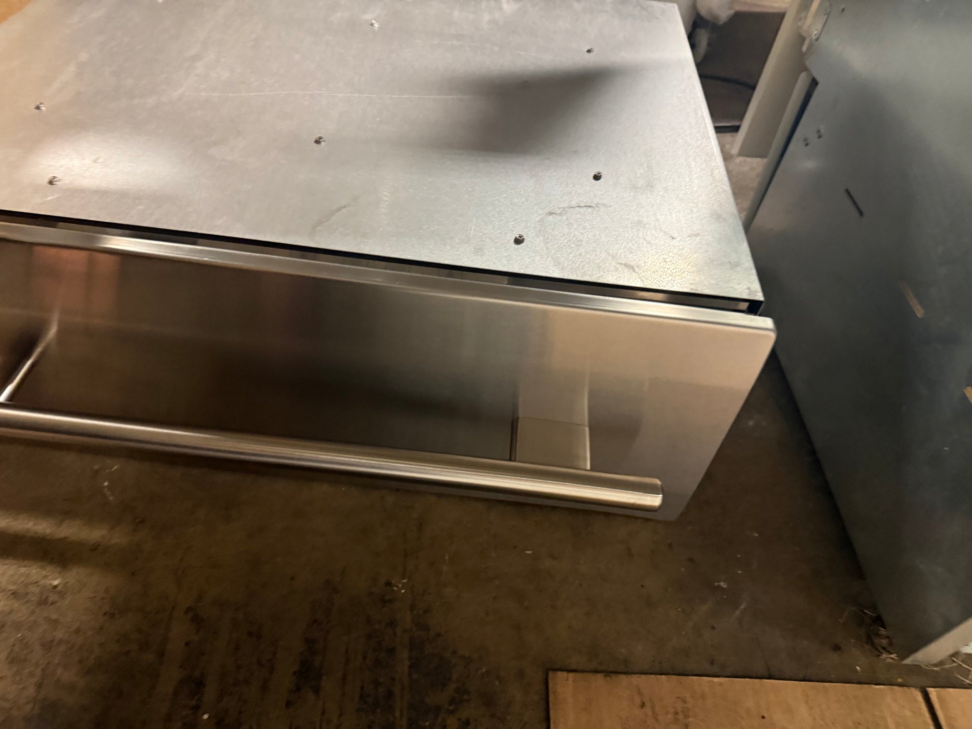 A stainless steel drawer-front appliance or cabinet component photographed in a warehouse-like setting, with a concrete floor and other metal objects visible around it.