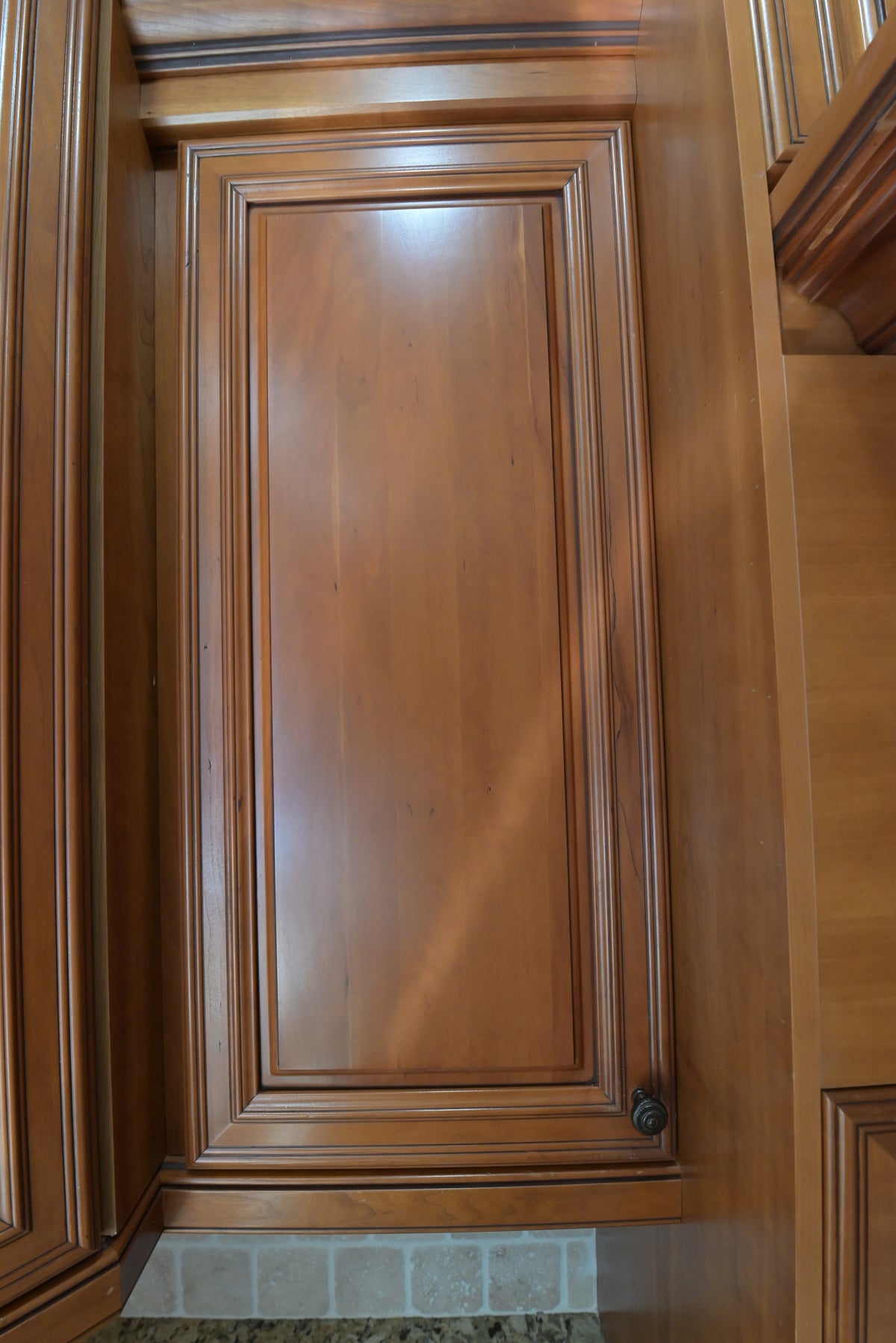 Aurora Circ traditional wood kitchen cabinet door with raised molding and dark knob above a tiled backsplash