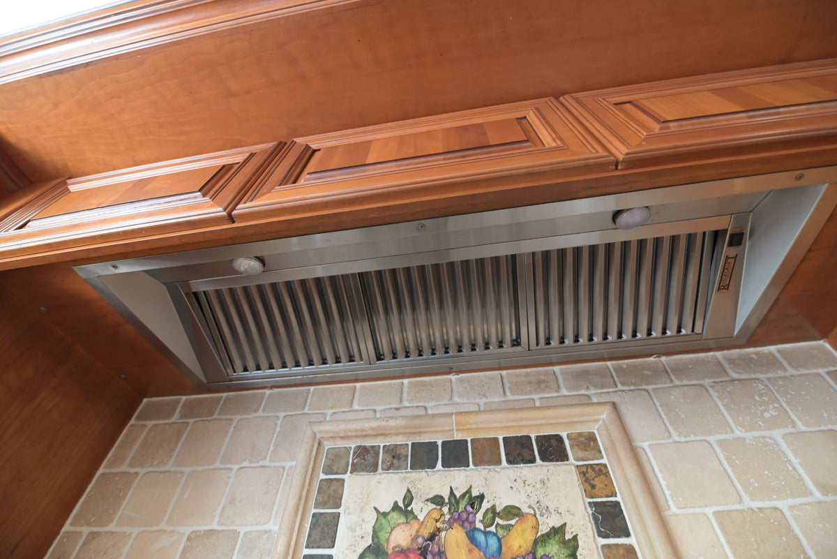 Aurora Circ traditional wood kitchen range hood with carved panel detailing above a tiled backsplash and decorative fruit mosaic