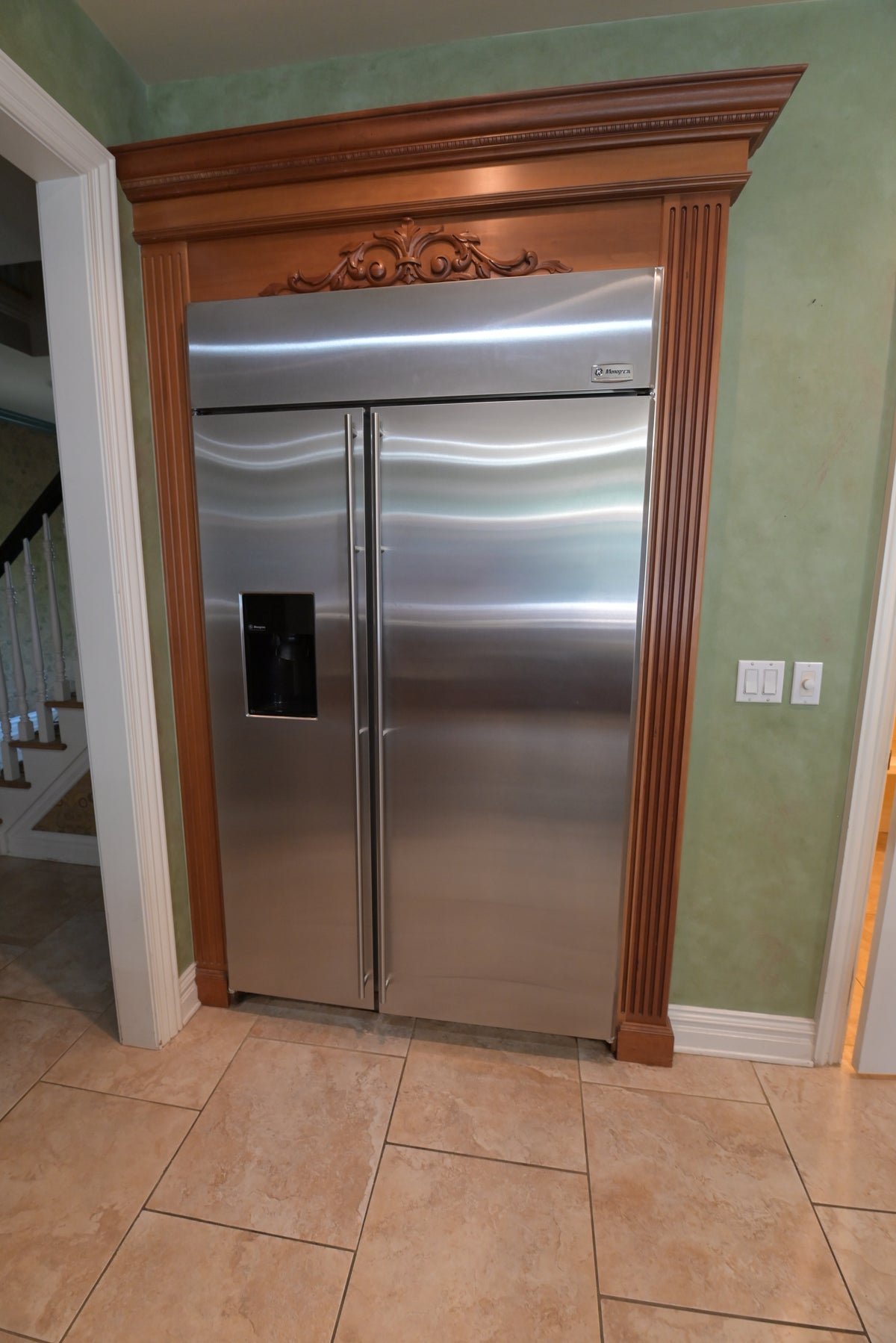 Aurora Circ stainless steel side by side refrigerator framed by ornate carved wood cabinetry and crown molding